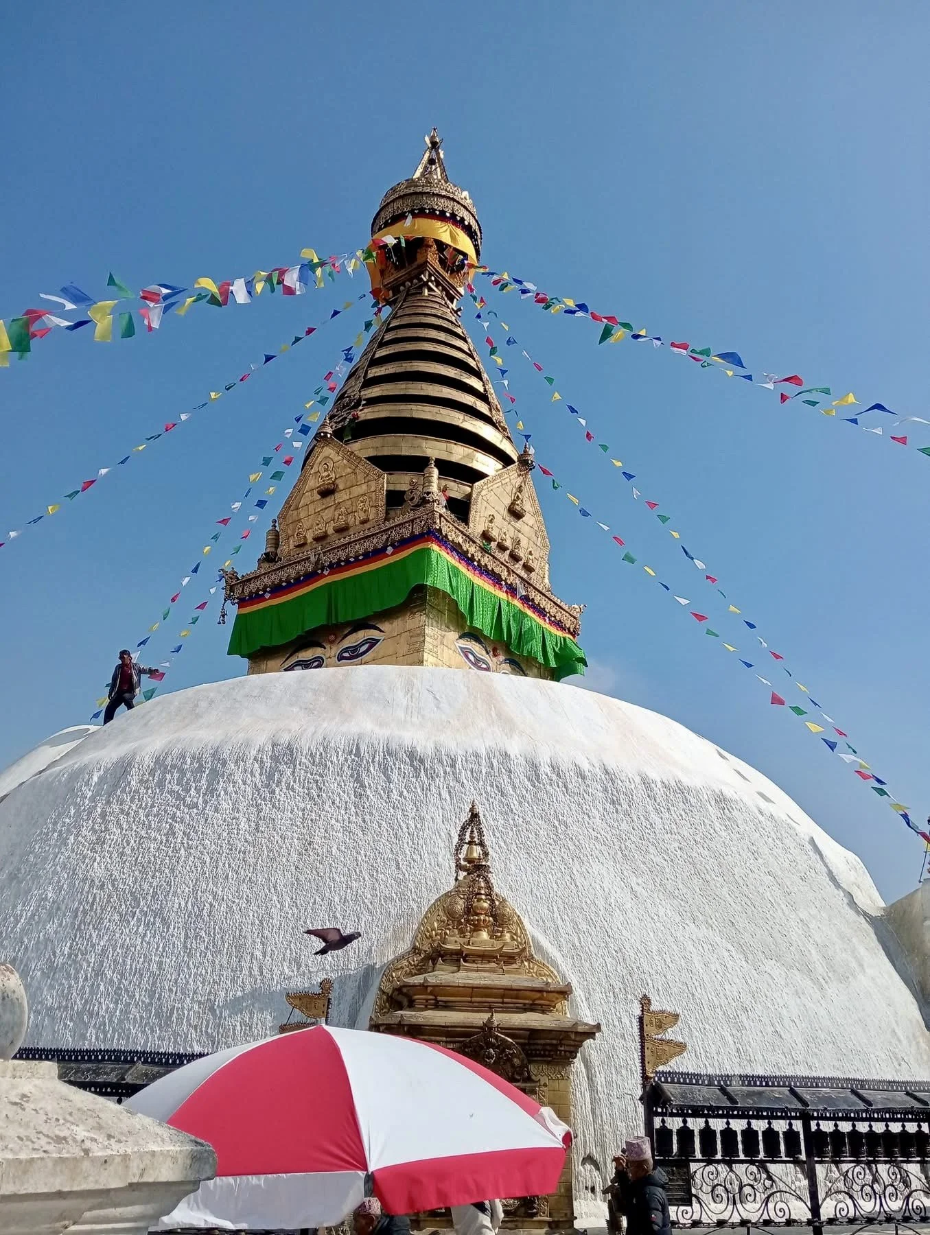 View of a white stupa or pagoda with a golden spire at the top, decorated with colorful prayer flags and a green cloth around its middle, set against a clear blue sky.