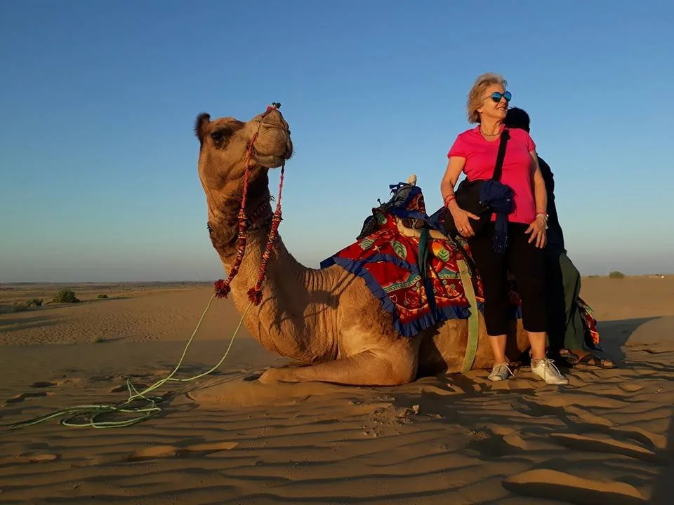 A woman standing beside a resting camel in a desert, with a clear sky in the background.