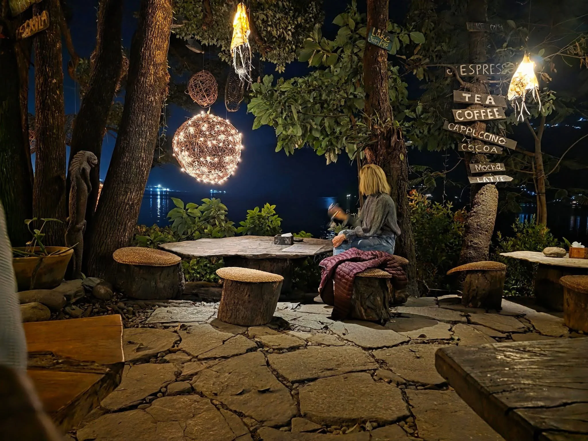 Night scene at an outdoor lakeside sit-down cafe with rustic wooden tables and stools, string lights, and leafy trees.