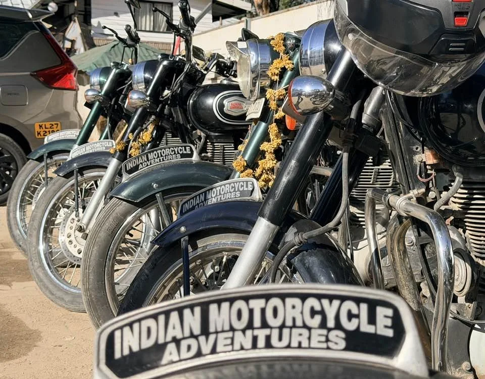 Multiple vintage Harley-Davidson motorcycles lined up outdoors, decorated with yellow marigold garlands, with a sticker reading 'Indian Motorcycle Adventures' on their fenders.