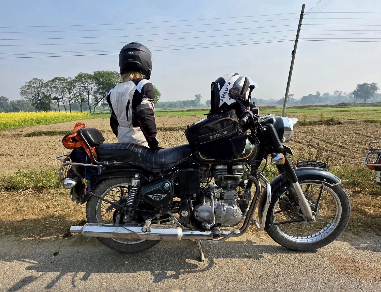 A person wearing a helmet and motorcycle gear standing next to a black vintage motorcycle on a rural road with fields and trees in the background.