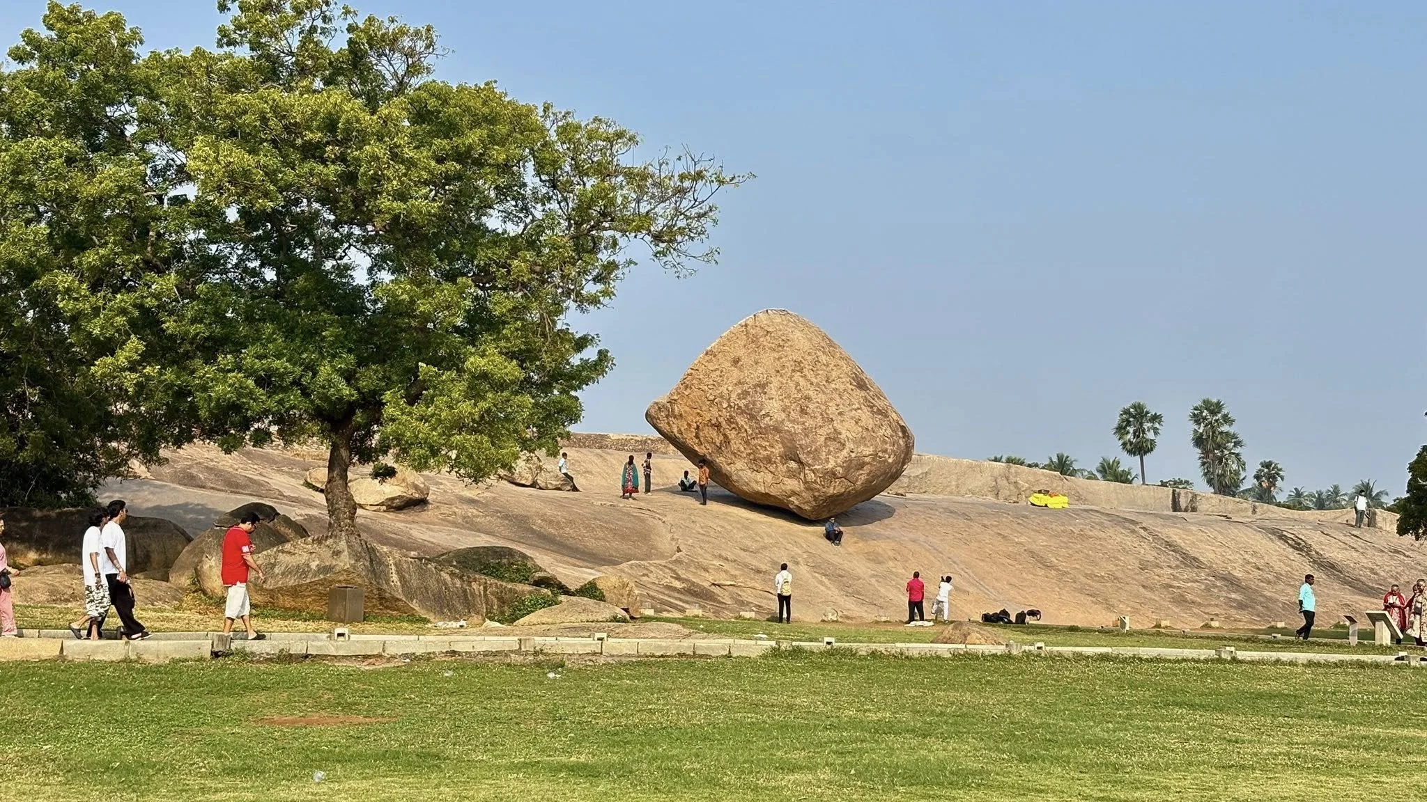 Large rock balanced on a sloped rocky surface with several people around it, some walking and some sitting, a green tree to the left, and palm trees in the background under a clear blue sky.