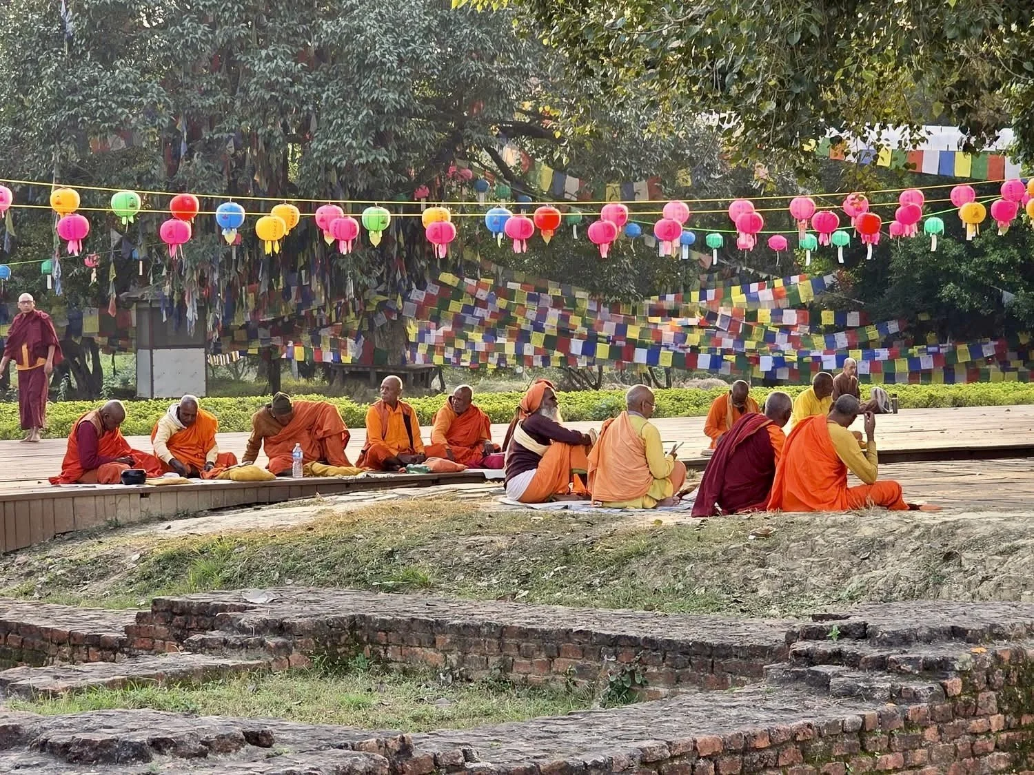 A group of monks, some dressed in orange, seated on a stage or platform outdoors, with colorful lanterns and prayer flags hanging overhead, surrounded by trees.