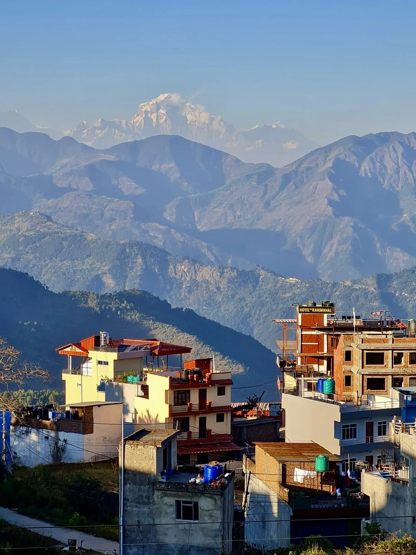 Hilly landscape with colorful buildings in the foreground, green hills, and a mountain range with snow-capped peaks in the background.