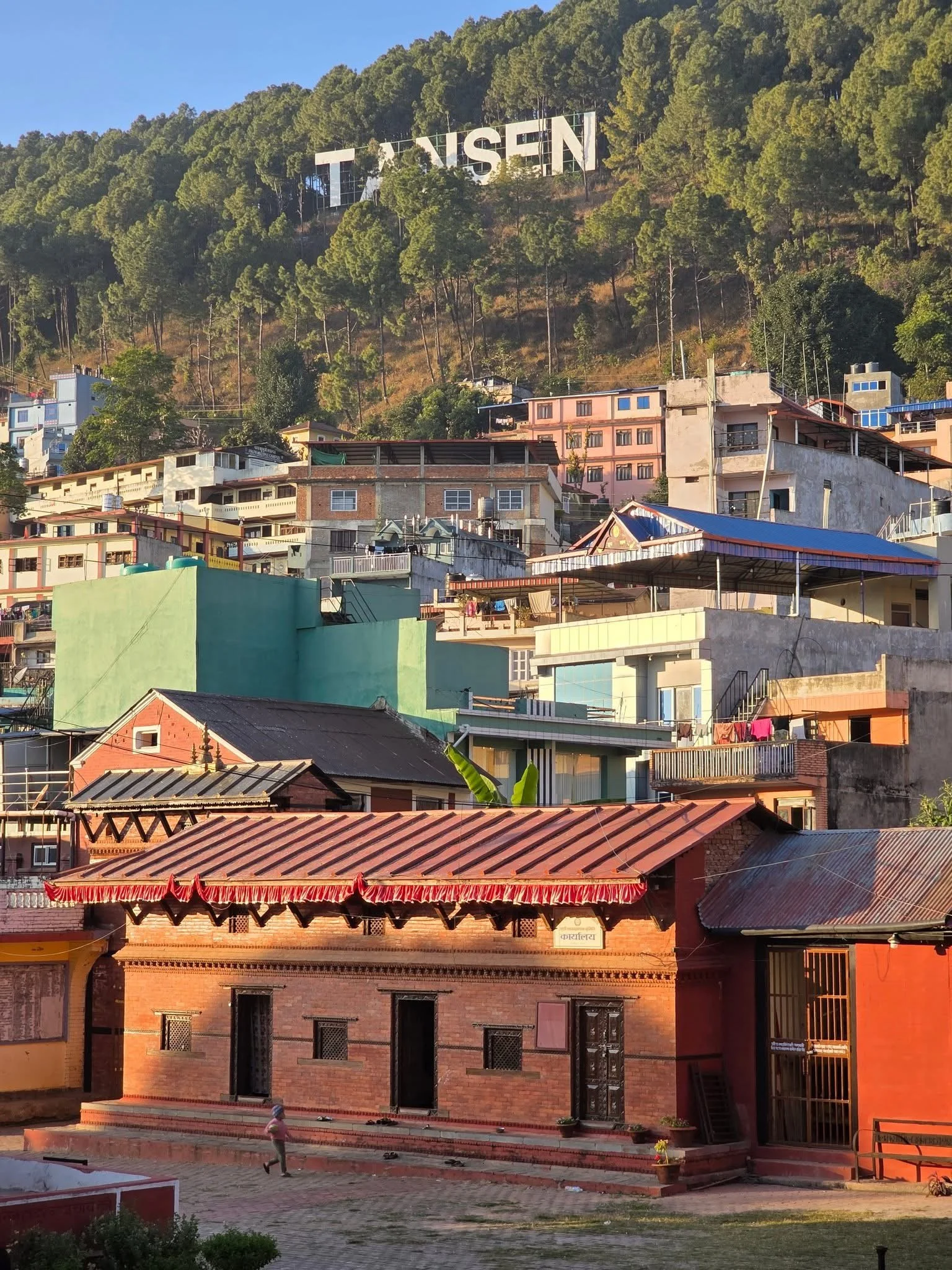 Colorful hillside town with various buildings, green trees, and a large hillside sign reading 'TANSEN' in the background.