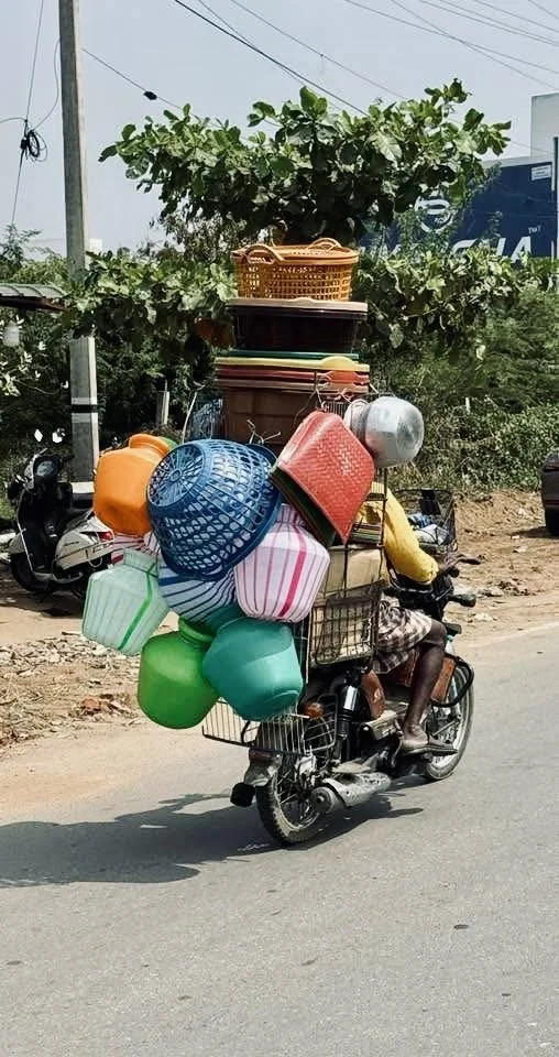 A man riding a motorbike with a large load of colorful household and shopping items, including buckets, baskets, and containers, stacked and tied to the bike on a street.