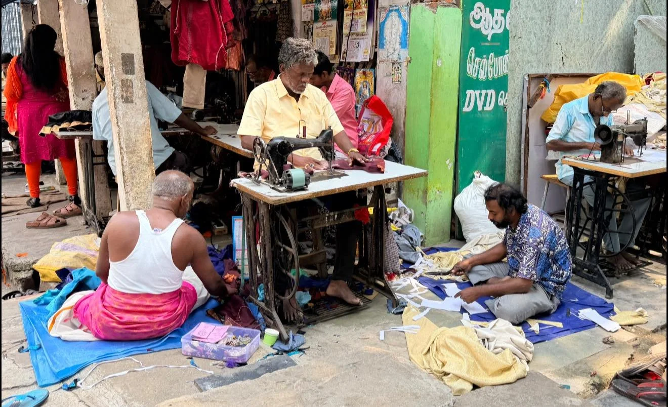 Street clothing workshop with multiple tailors sewing fabrics and garments outside shops in an urban setting.