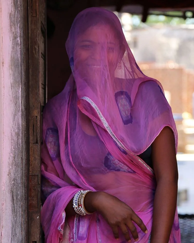 A woman wearing a pink and purple sari with a sheer veil over her head, smiling, and wearing colorful bangles on her wrist.