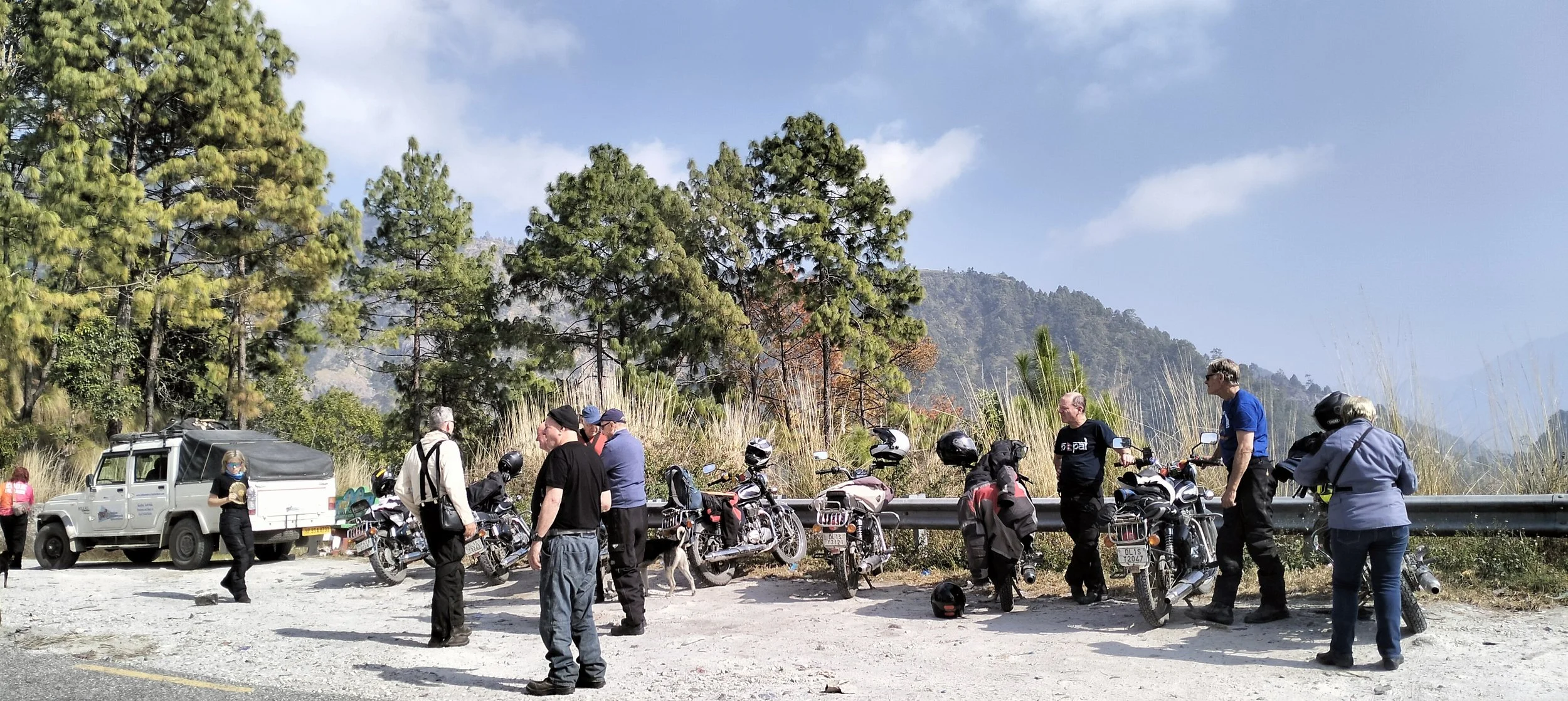 Group of motorcyclists and a police officer gathered in a scenic mountain area with trees and blue sky.
