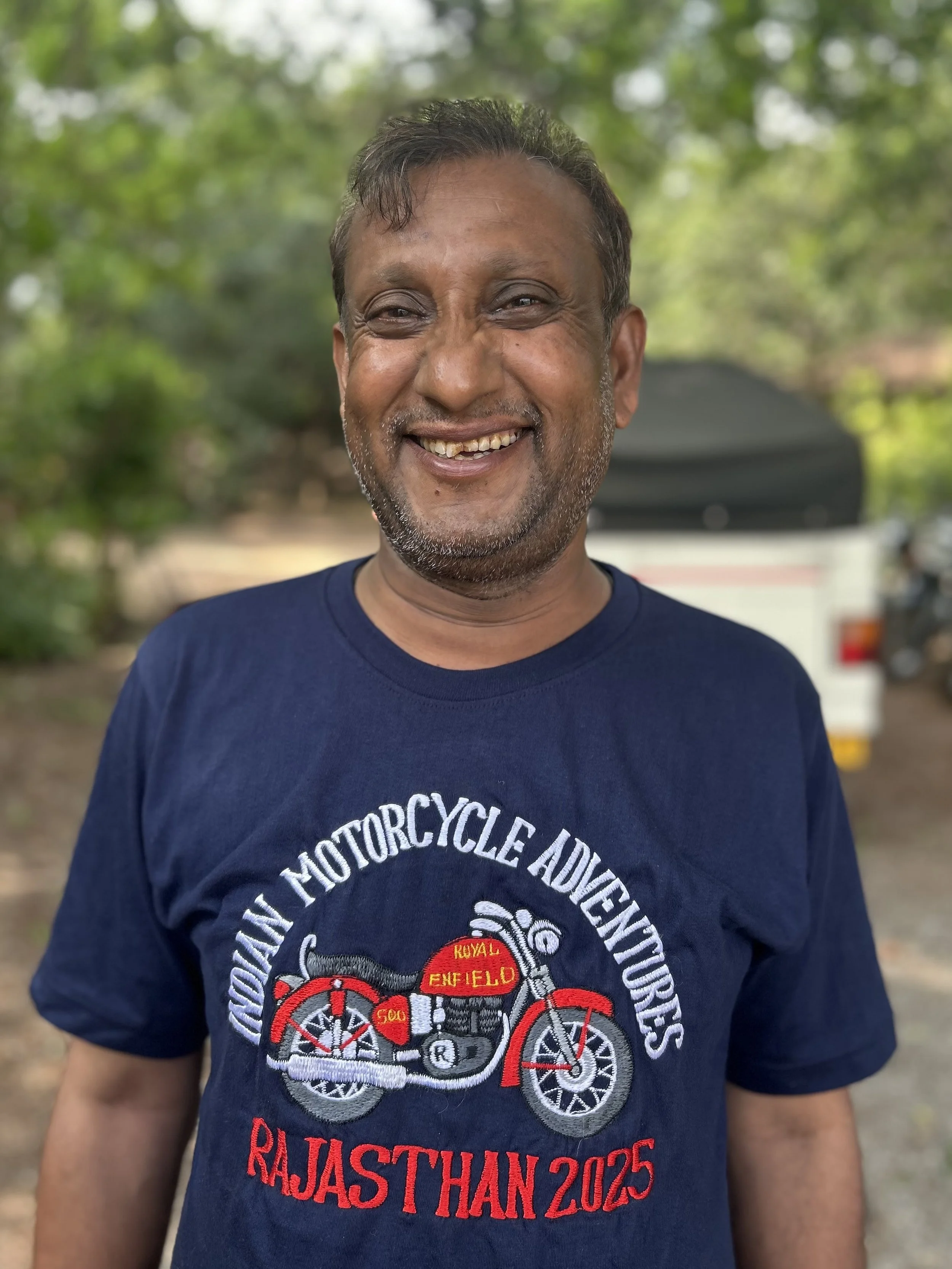 A man in a black and white striped shirt leaning on a motorcycle on a street, with a colorful bus and trees in the background.
