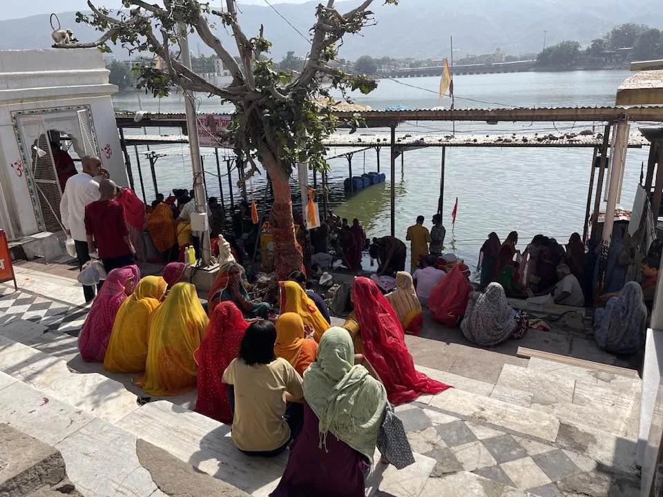 A group of women wearing colorful traditional clothing sits on marble steps near a river, some are praying or meditating, with others standing or walking nearby under a large tree. In the background, there are more people near the water and structure