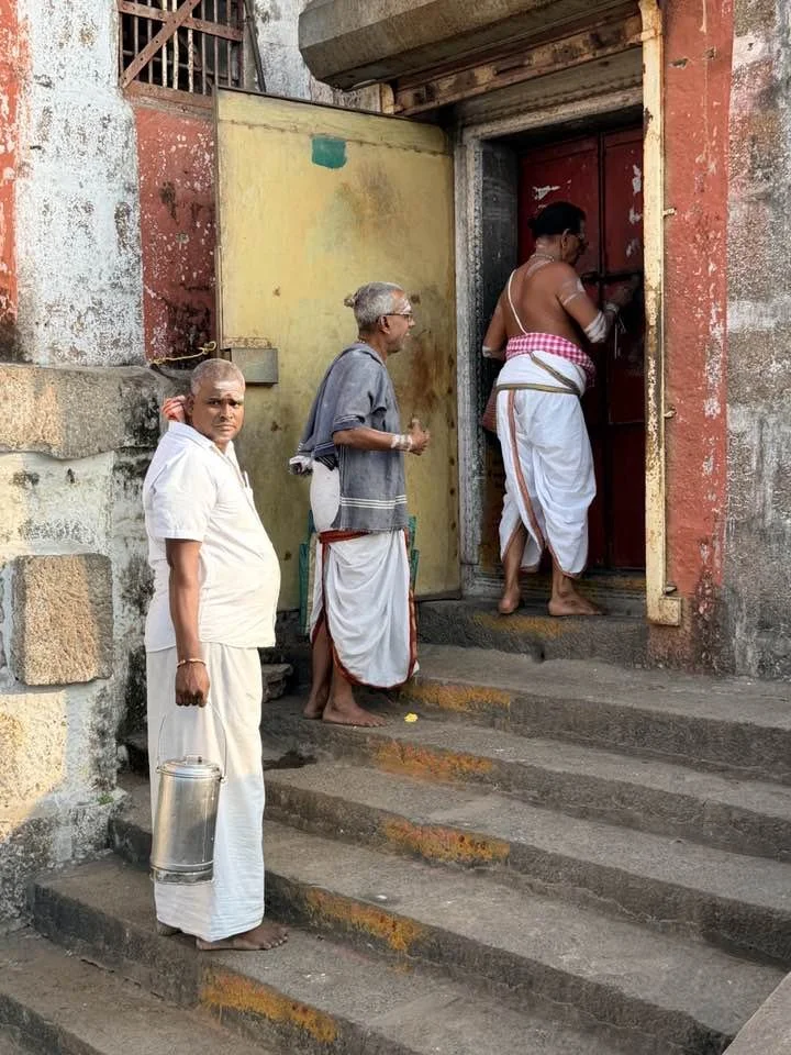 Three men are standing on steps outside a building, one is in the process of opening a red door. The men are dressed in traditional Indian attire, with two wearing white dhotis and one wearing a grey kurta. One man is holding a metal container.