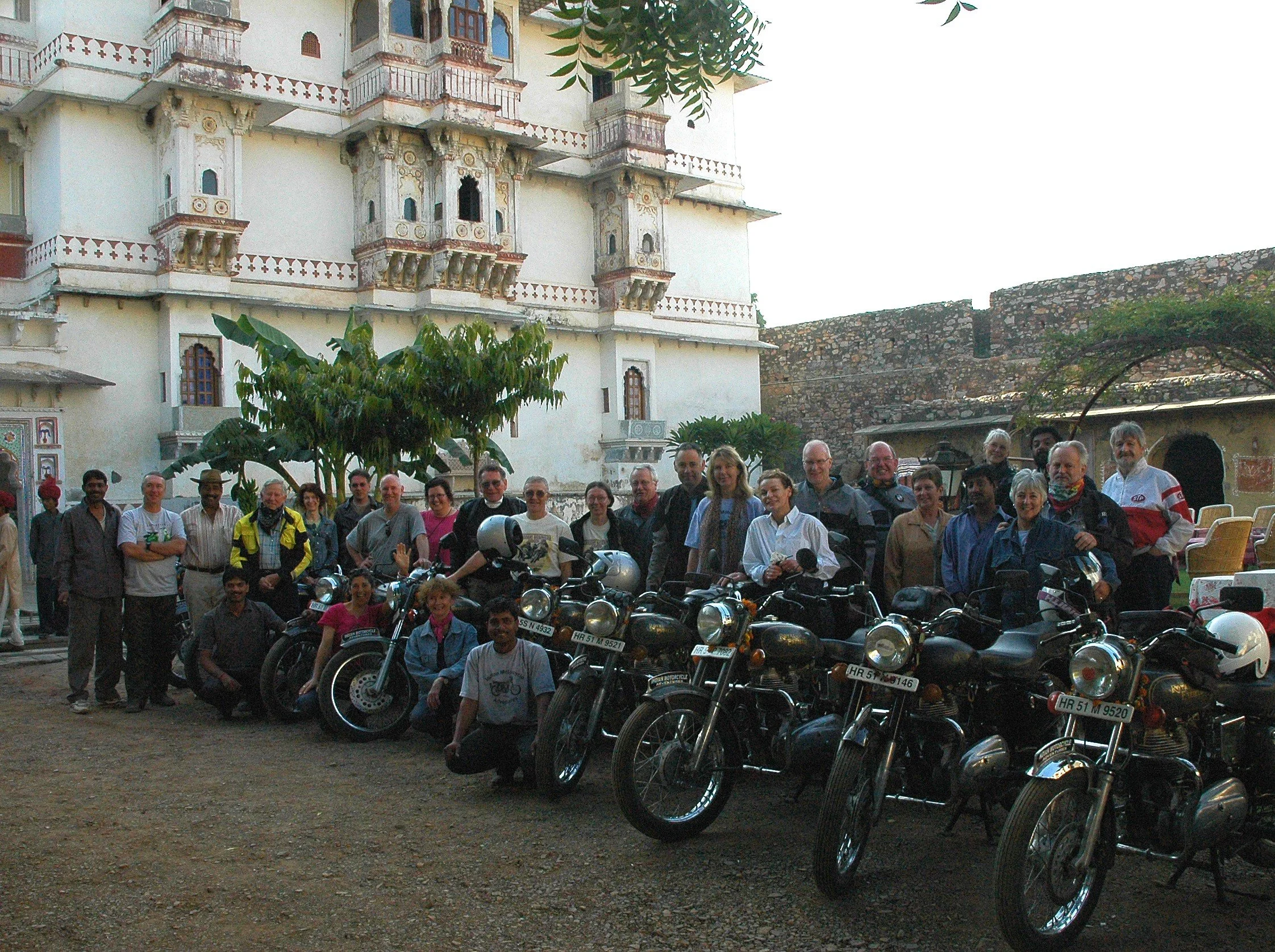 A group of people standing and sitting in front of a row of motorcycles, with a historic building in the background.