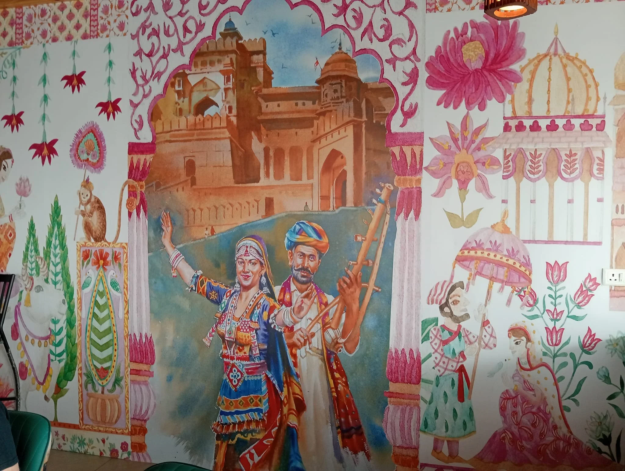 Colorful mural depicting traditional Indian dancers in vibrant clothing, floral motifs, and architectural elements, including a castle and temple structures, with bright pink, green, and gold accents.