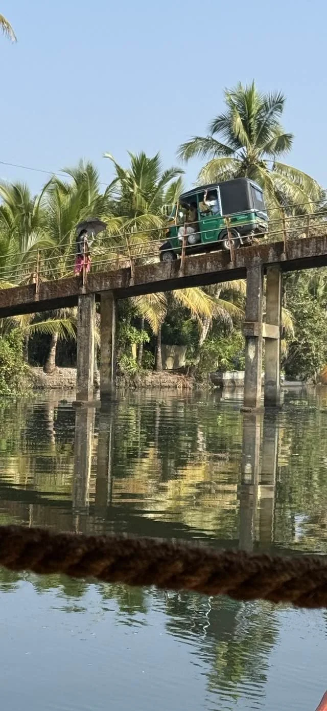 A small green auto rickshaw crossing a bridge over a canal, with a person holding an umbrella walking nearby and palm trees in the background.