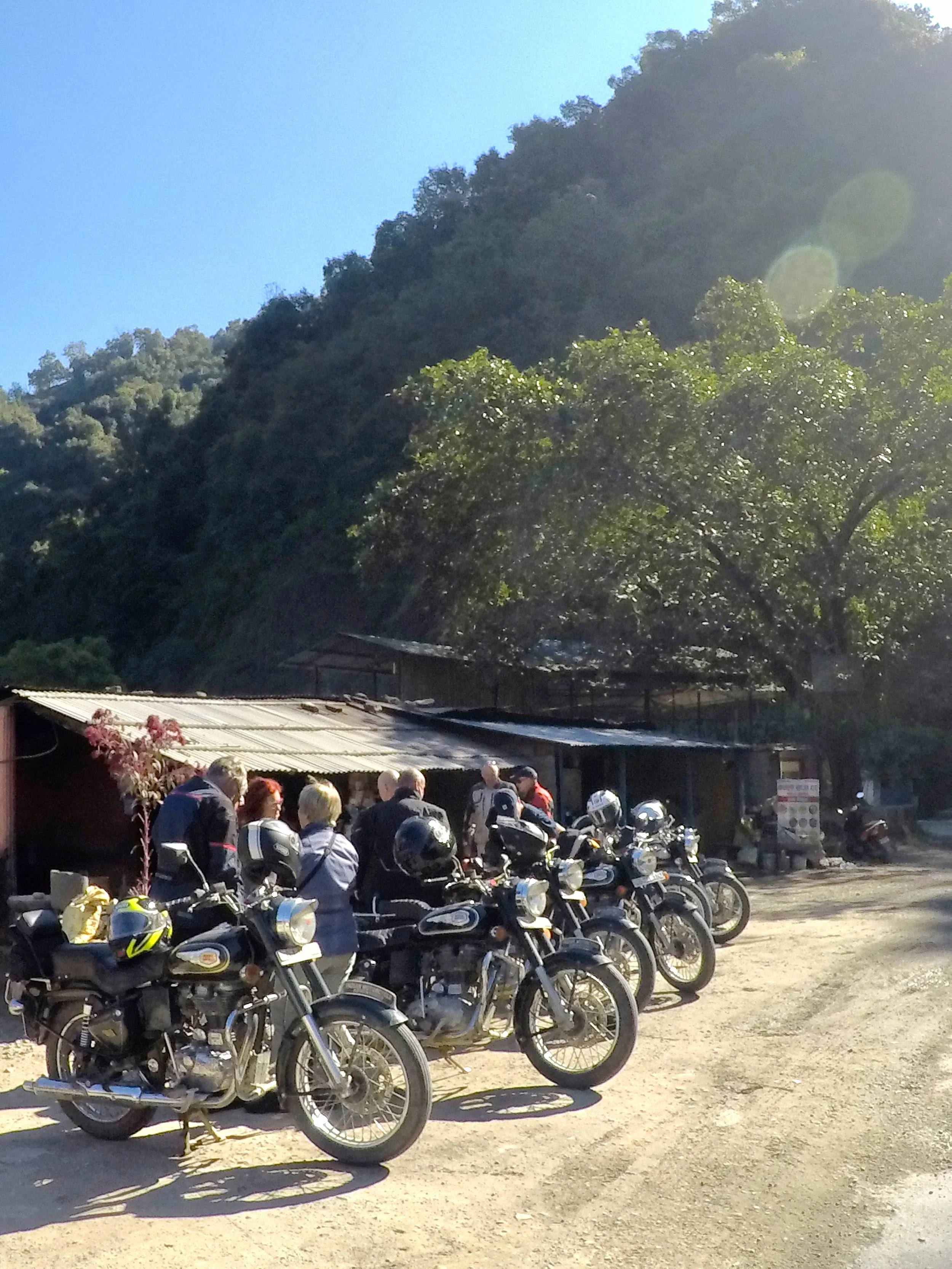 Group of motorcyclists and passengers with helmets, parked on a dirt roadside near a rustic building, with a tree and mountain in the background, sunny weather.