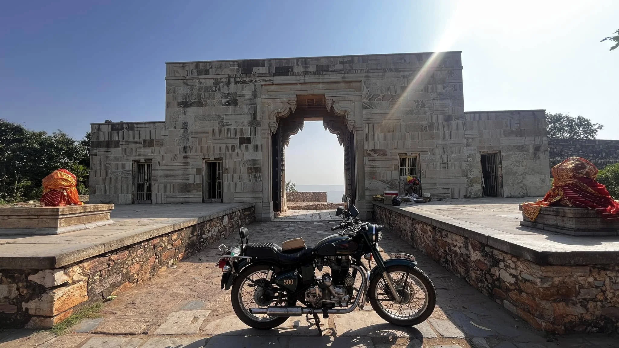 A black vintage motorcycle with a brown seat parked in front of an ancient stone temple with arched entrance, small windows, and decorative elements, under a bright blue sky with sunlight.