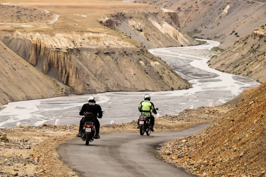 Two motorcyclists riding on a narrow mountainous road overlooking a winding river in a rugged, arid landscape.