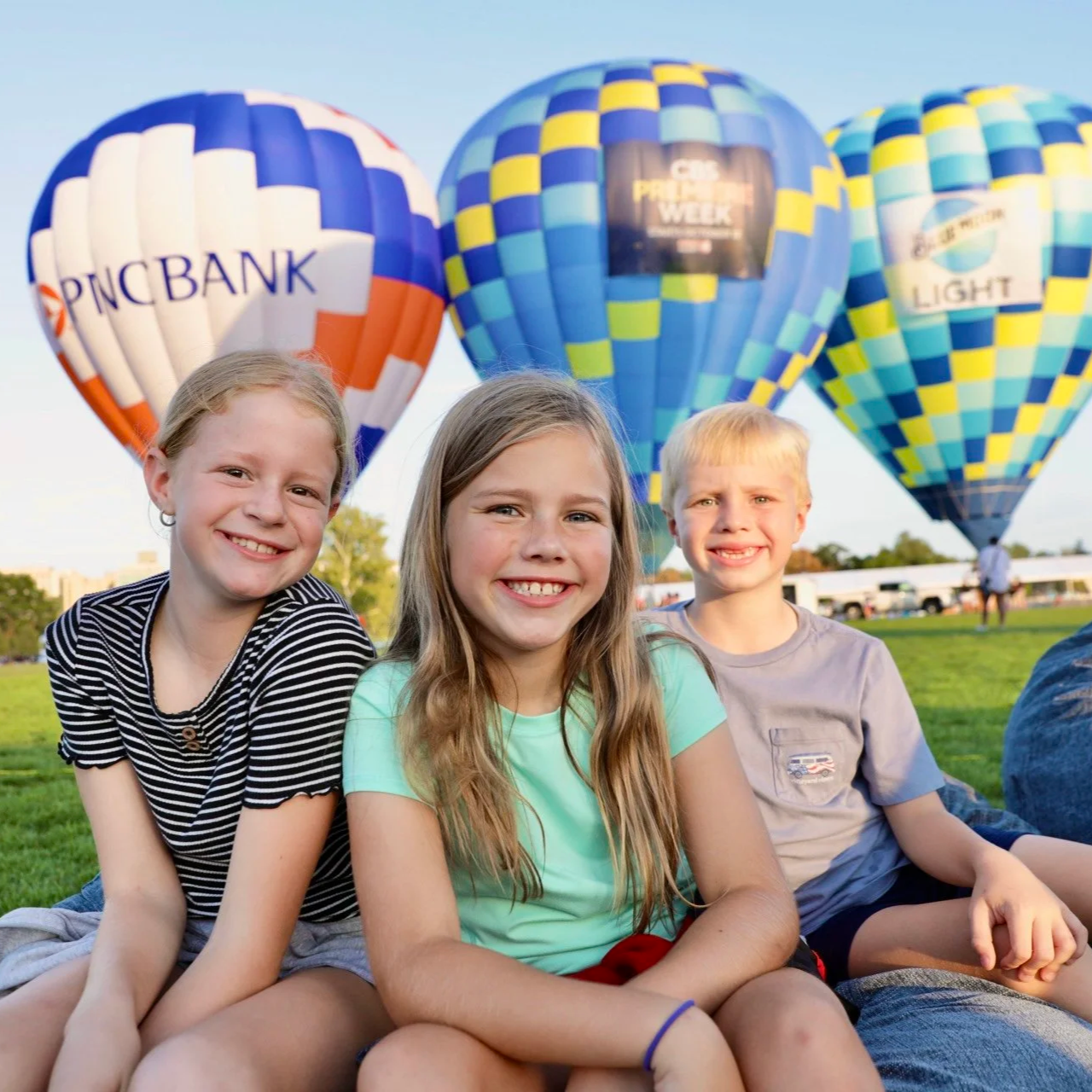 Three children sitting on grass at a hot air balloon festival with three colorful hot air balloons in the background.