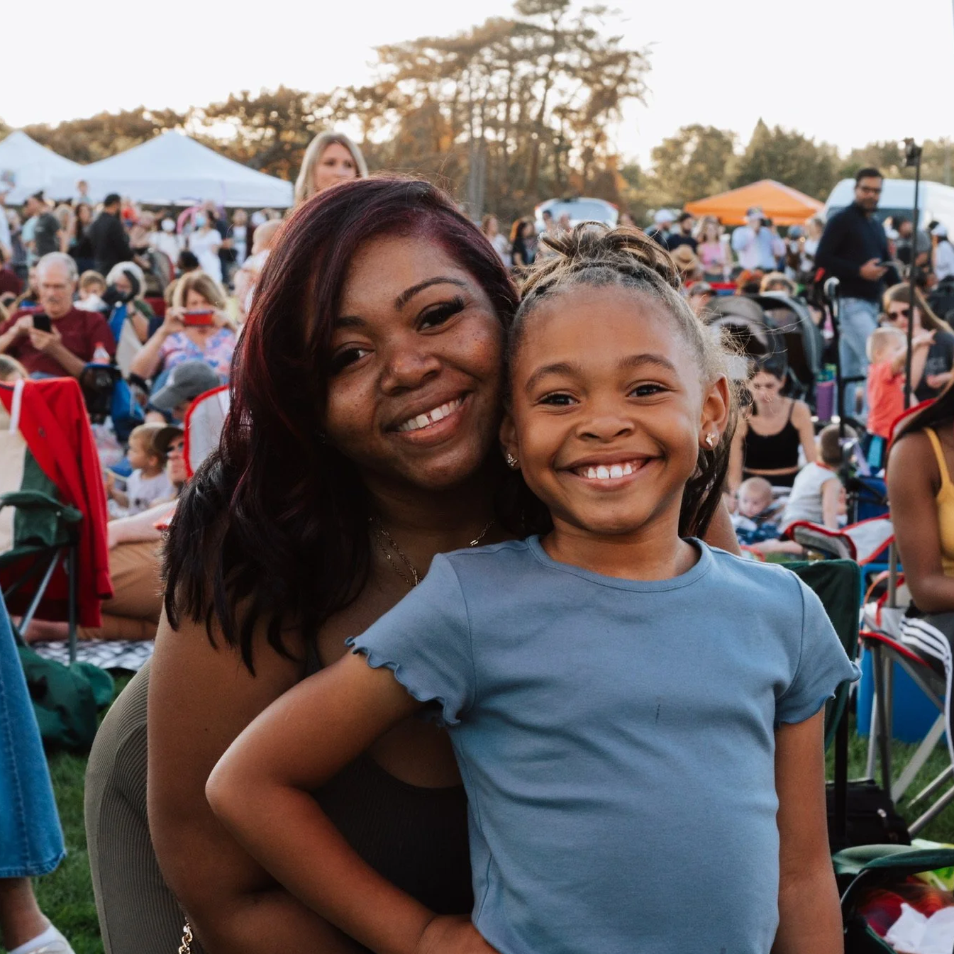 A woman and a little girl smiling at an outdoor event with tents and many people in the background.