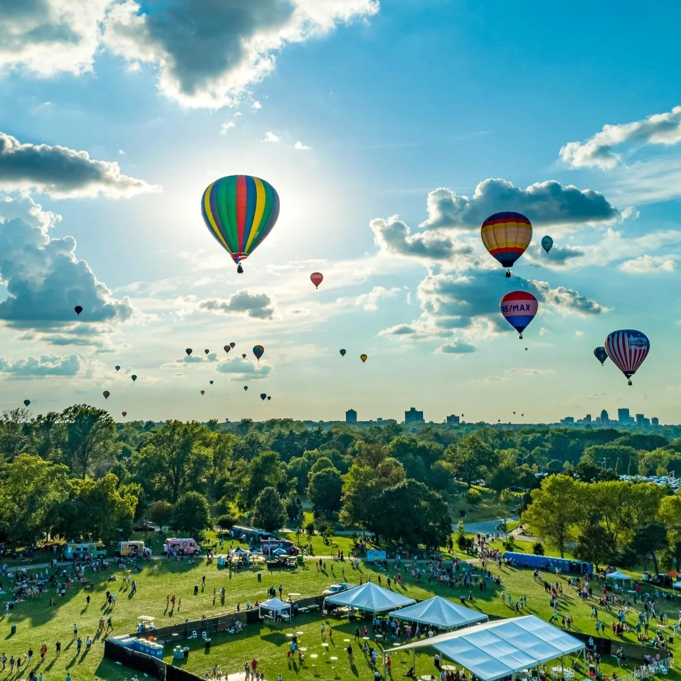 Multiple colorful hot air balloons flying in a partly cloudy sky over a park with trees and a crowd of people, tents, and vehicles on the ground.