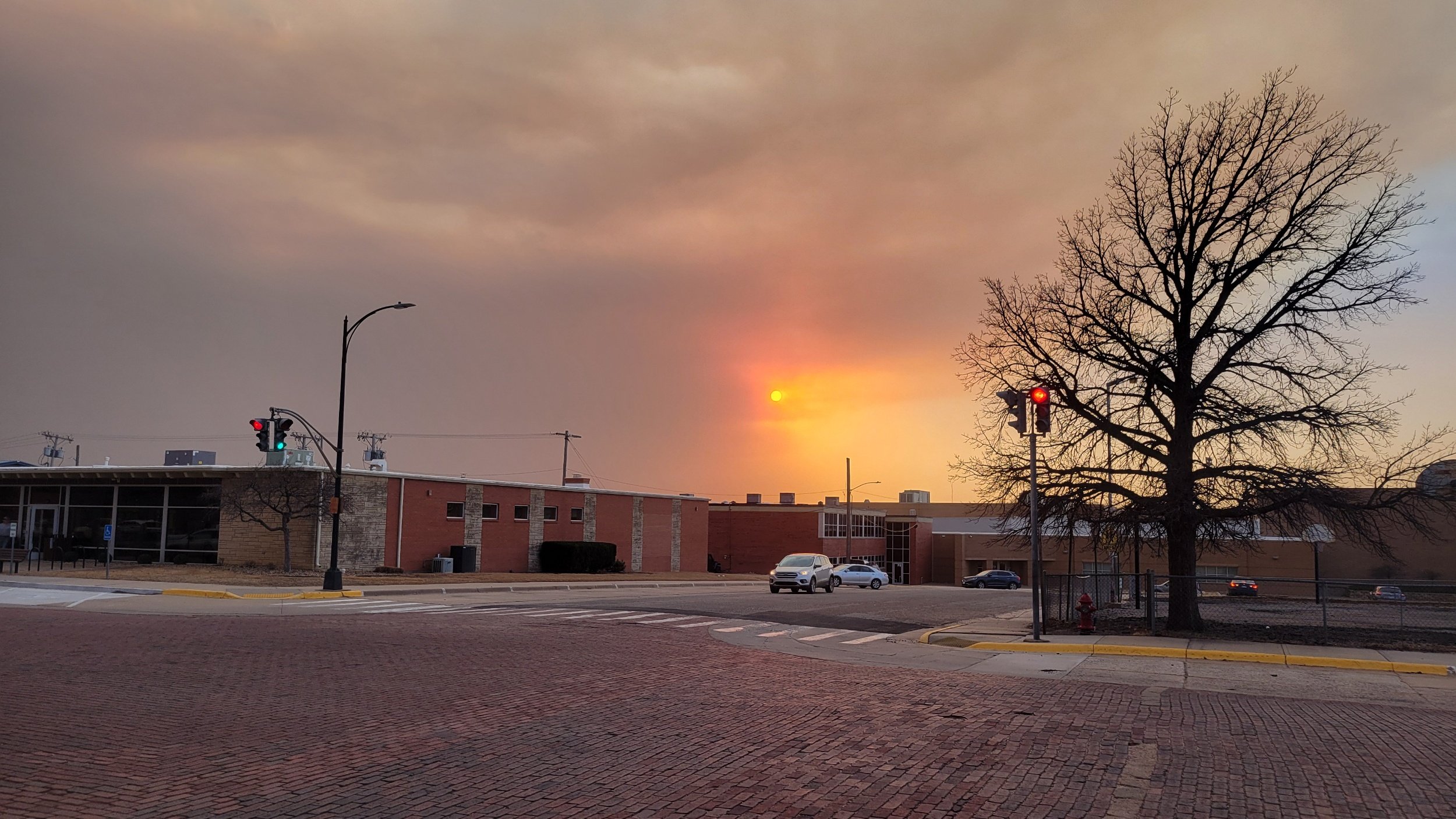 City intersection with traffic lights, a leafless tree, and buildings during sunset with a cloudy sky.