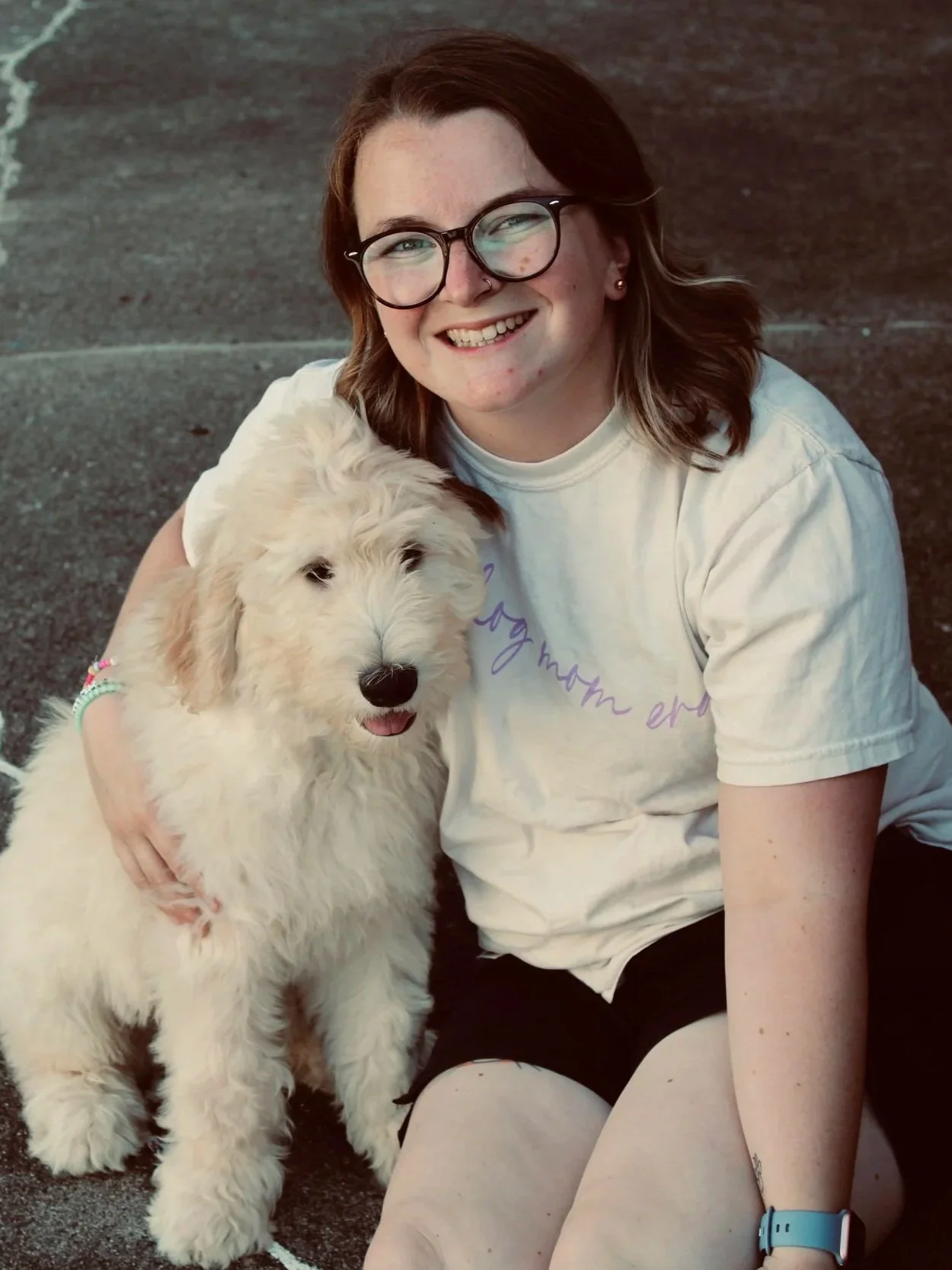 A young woman with glasses and long brown hair smiling while sitting on the ground and hugging a large fluffy white dog with floppy ears and a happy expression.