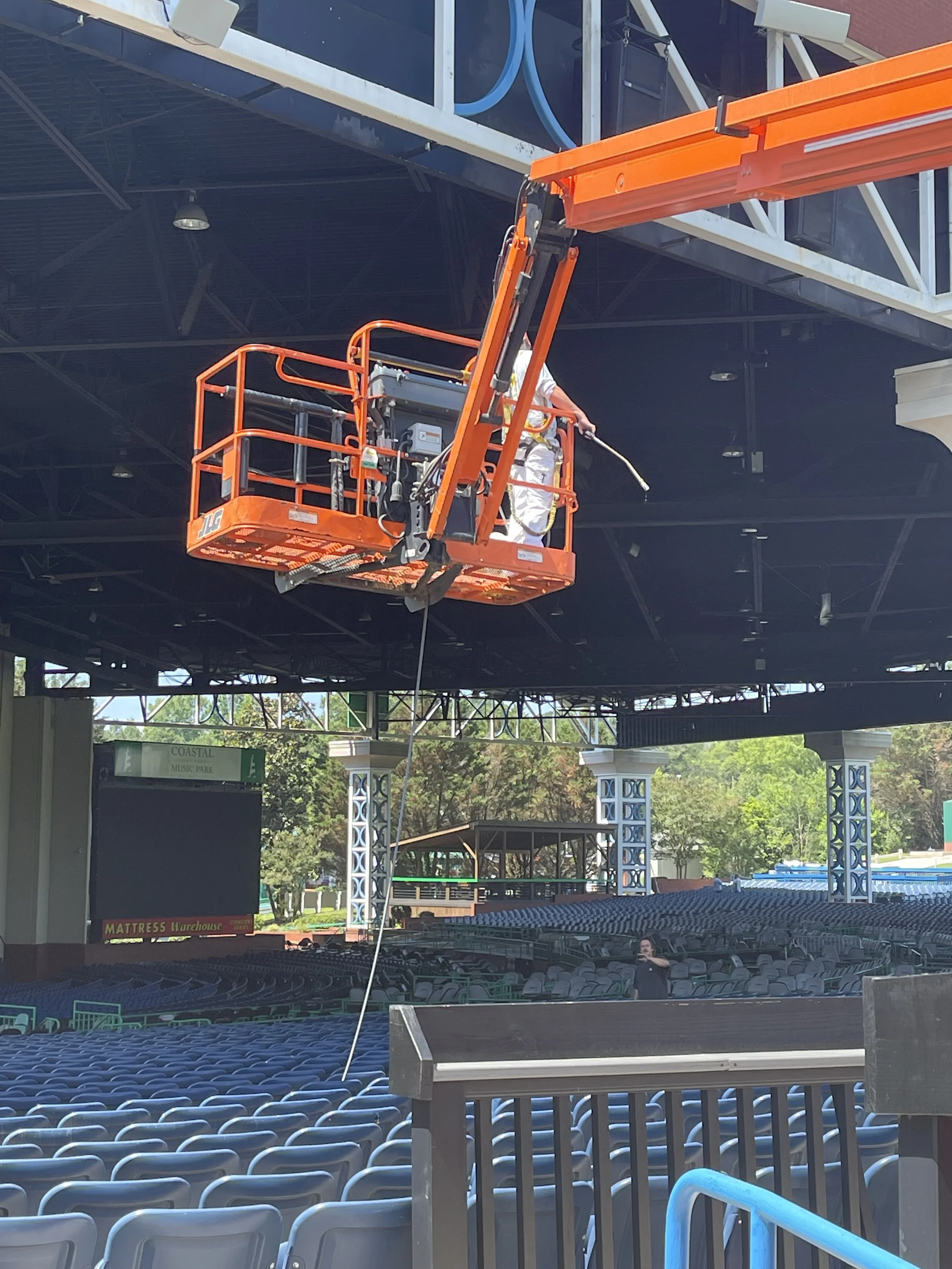 A worker standing on an orange lift is cleaning or inspecting the stage ceiling of an outdoor theater while an empty audience area sits below.
