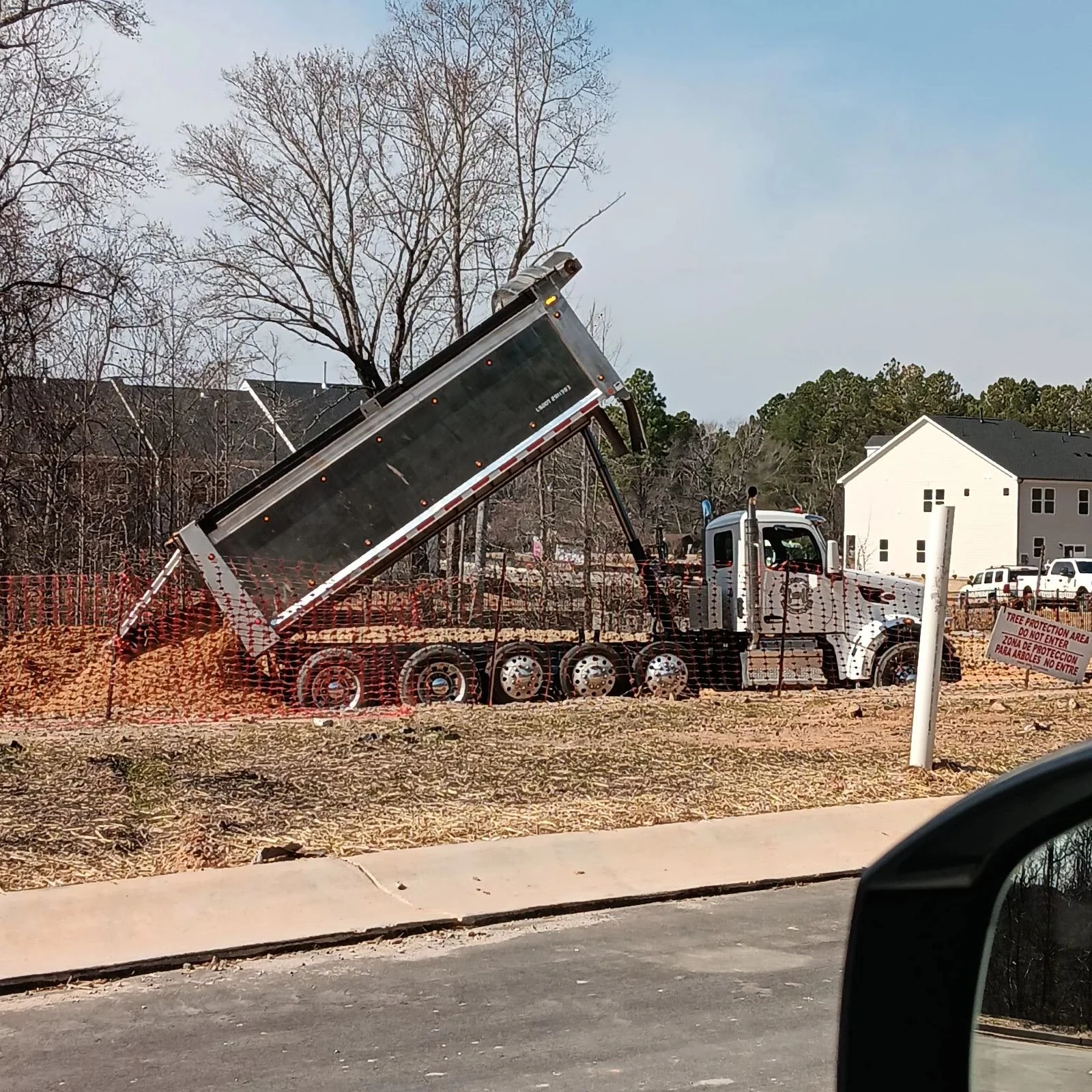Construction site with a dump truck tilted to unload dirt, surrounded by orange safety barrier fencing, near a residential area with trees and houses in the background.