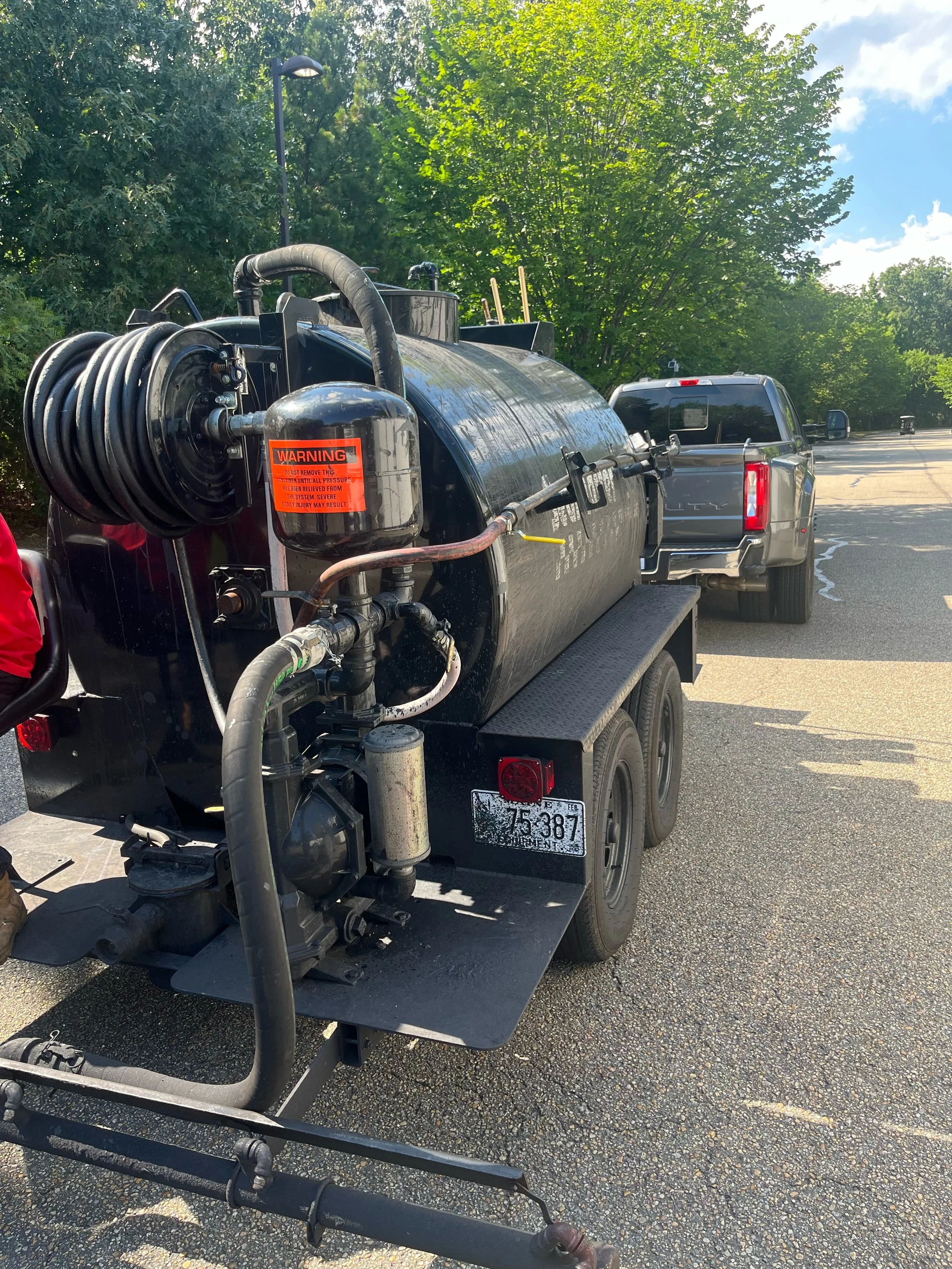 Black tanker trailer with hoses and warning labels parked on a street with trees and blue sky in the background.