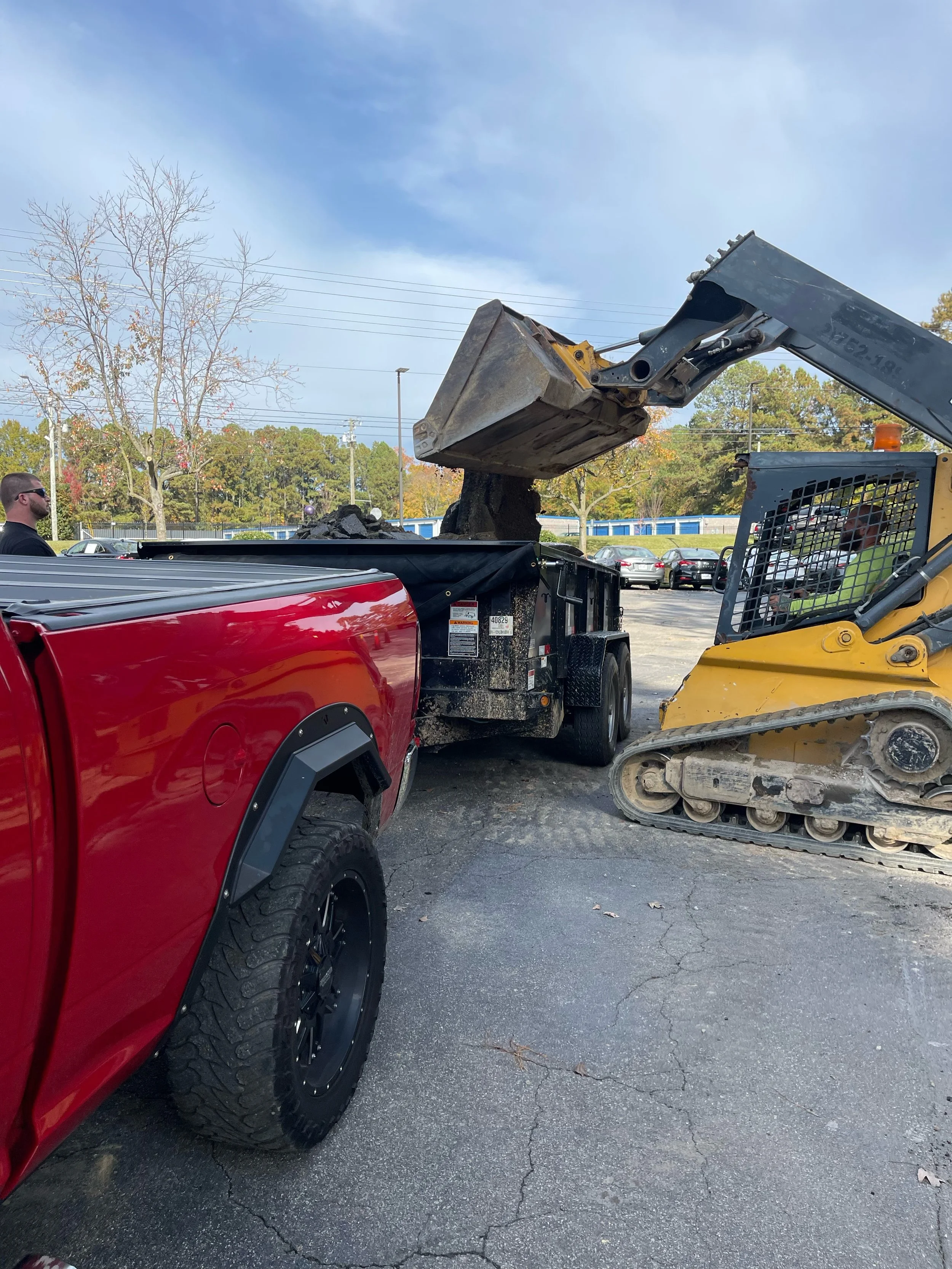 A yellow mini excavator unloading dirt into the bed of a red pickup truck in a parking lot.