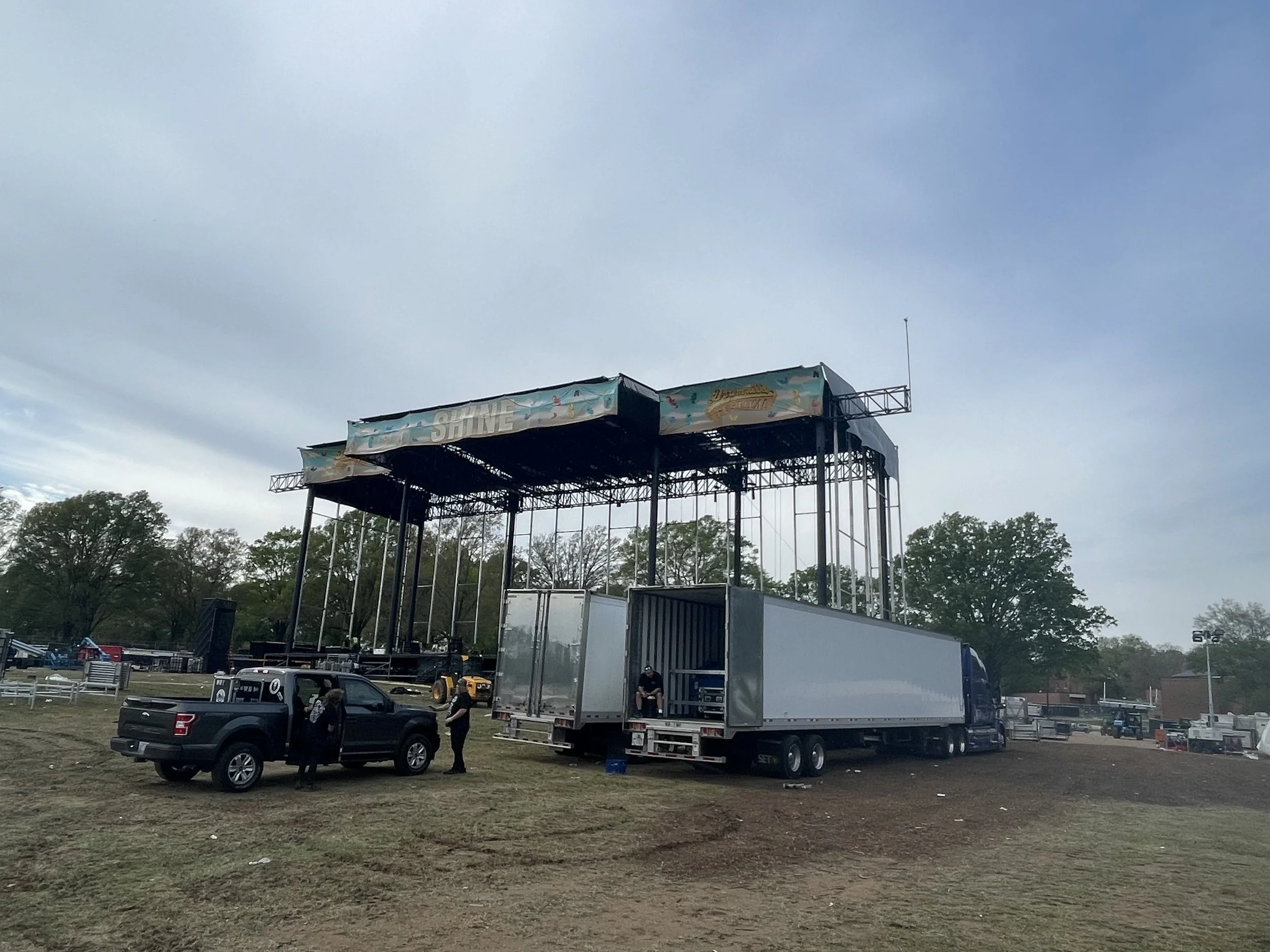 Stage being assembled outdoors with trucks and equipment, trees in the background, cloudy sky overhead.