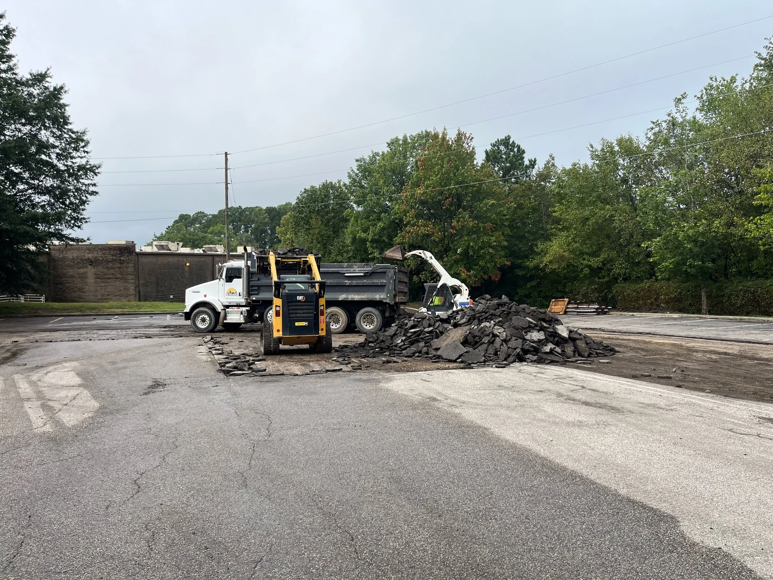 Construction workers repair a damaged road with asphalt and rocks using heavy machinery during daytime, surrounded by trees and overcast sky.