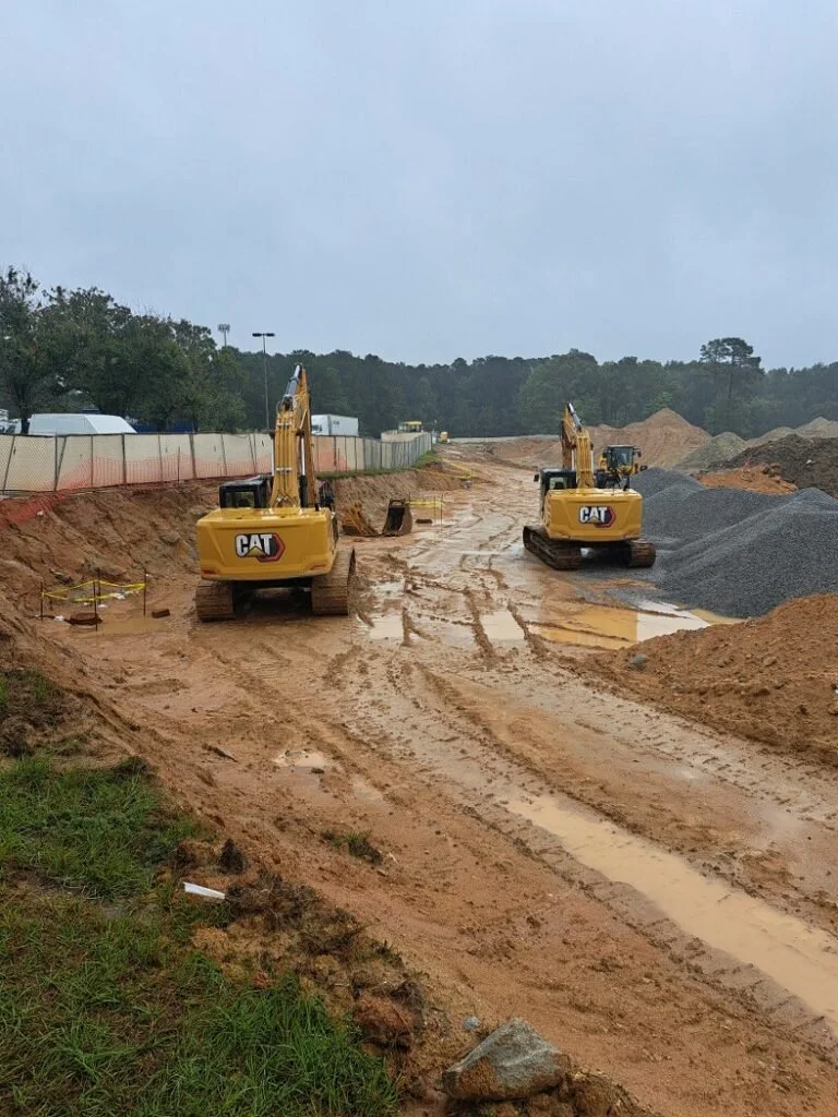 Construction site with two yellow Caterpillar excavators working on muddy terrain, piles of dirt and gravel, with trees and overcast sky in the background.