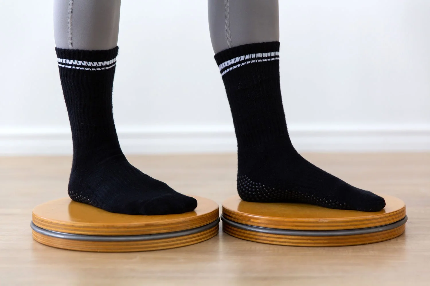 Side view of a person standing on wooden Pilates rotating discs, wearing black grip socks and grey leggings.