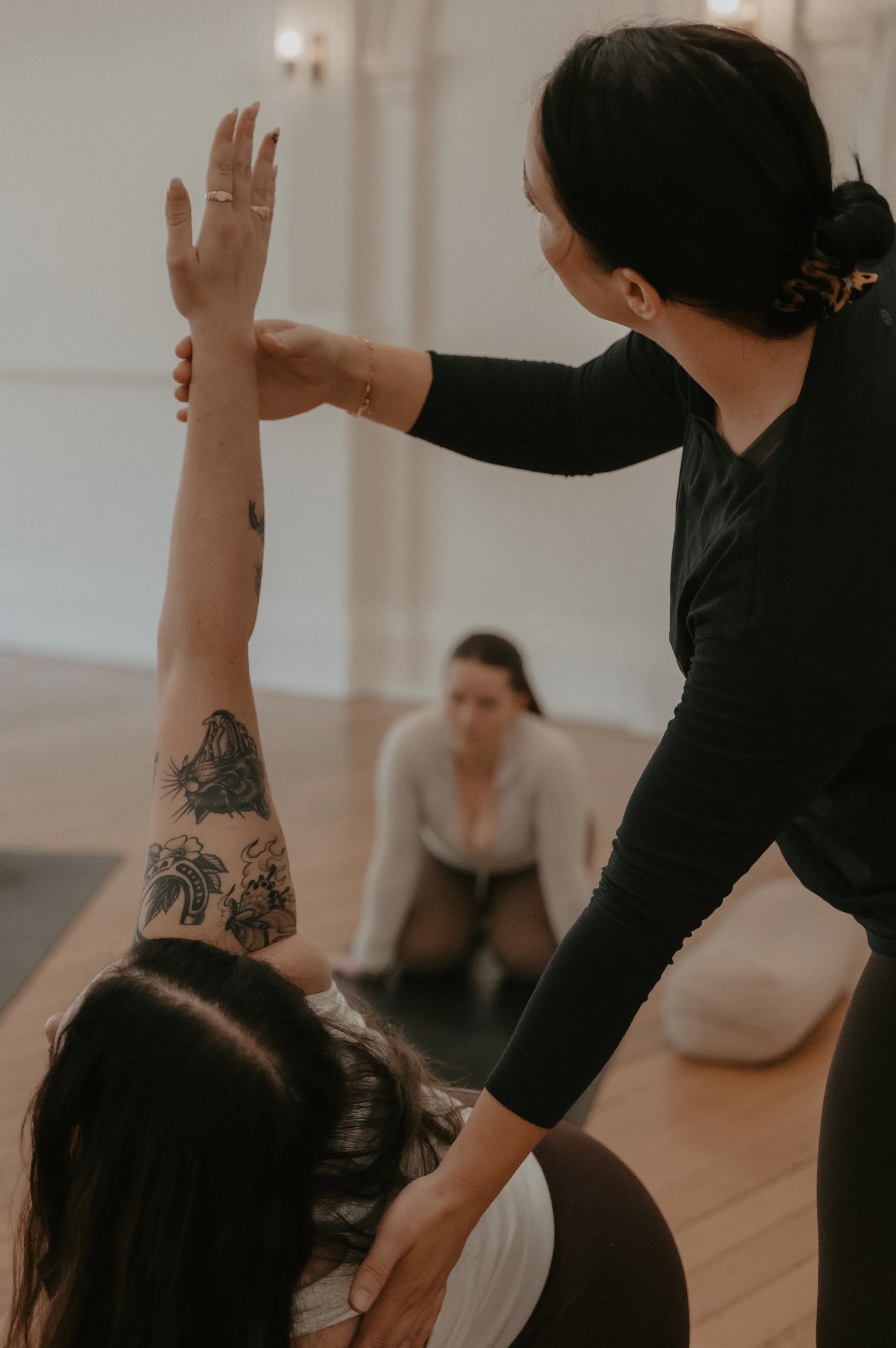 Pilates instructor assisting a woman with pilates pose during pilates teacher training, with another woman practicing in the background.