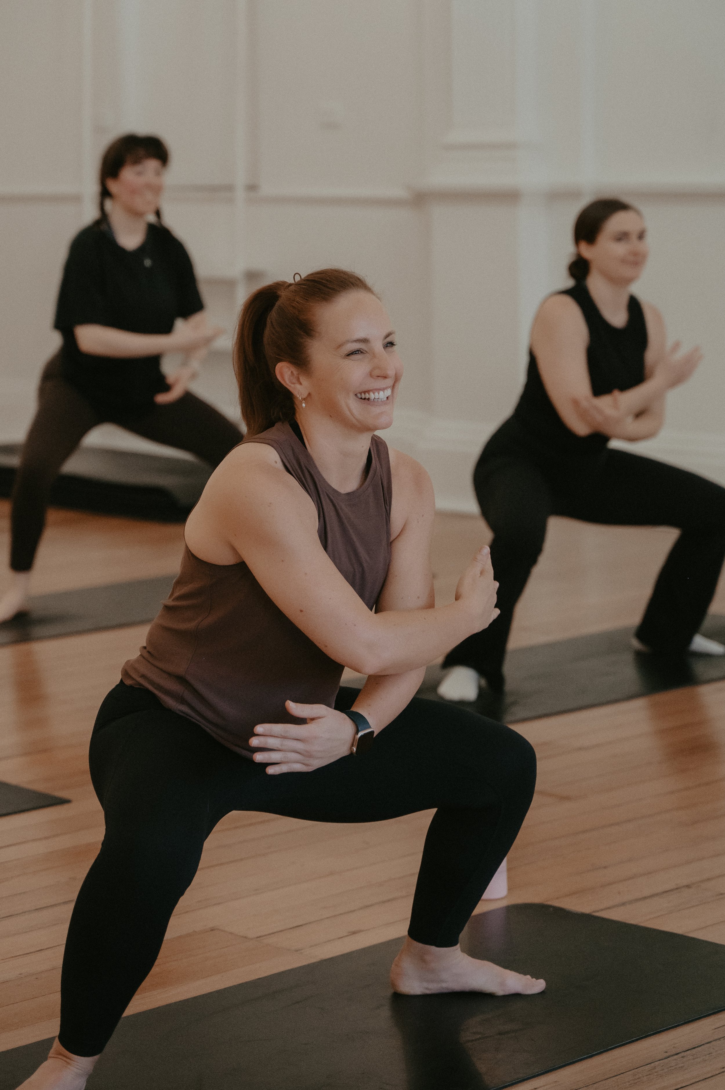 Women participating in a pilates class in a room with wooden floors, practicing pilates poses and smiling.