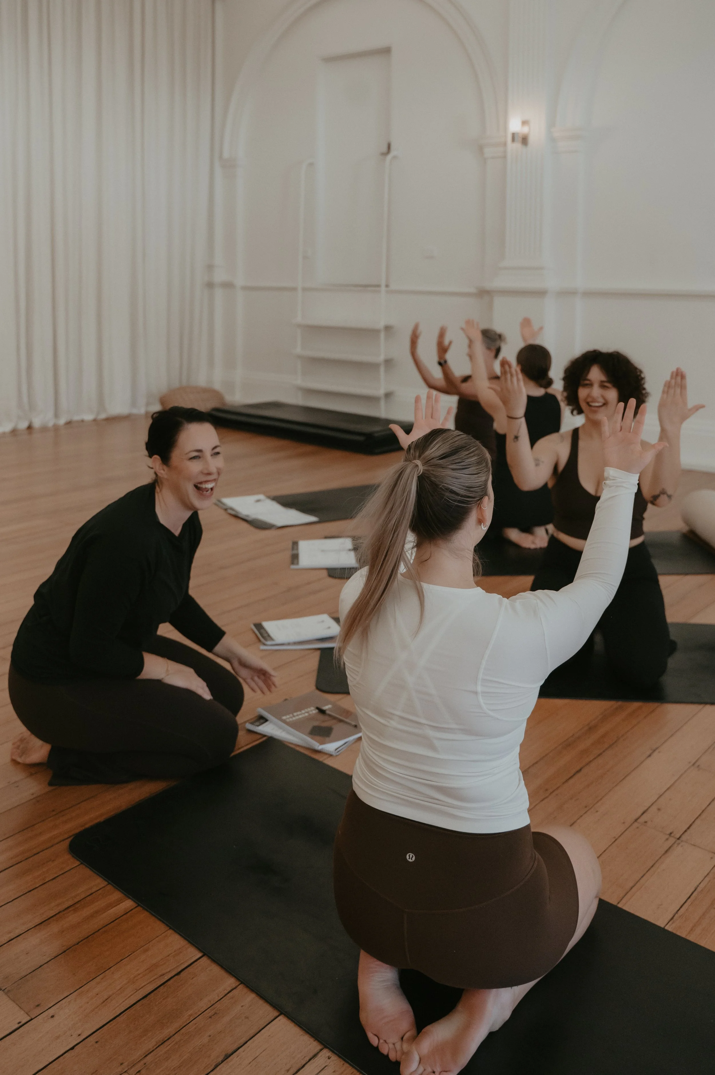 A group of women participating in a fitness or pilates class in a studio, sitting on black mats with their arms raised, smiling and engaging with each other.