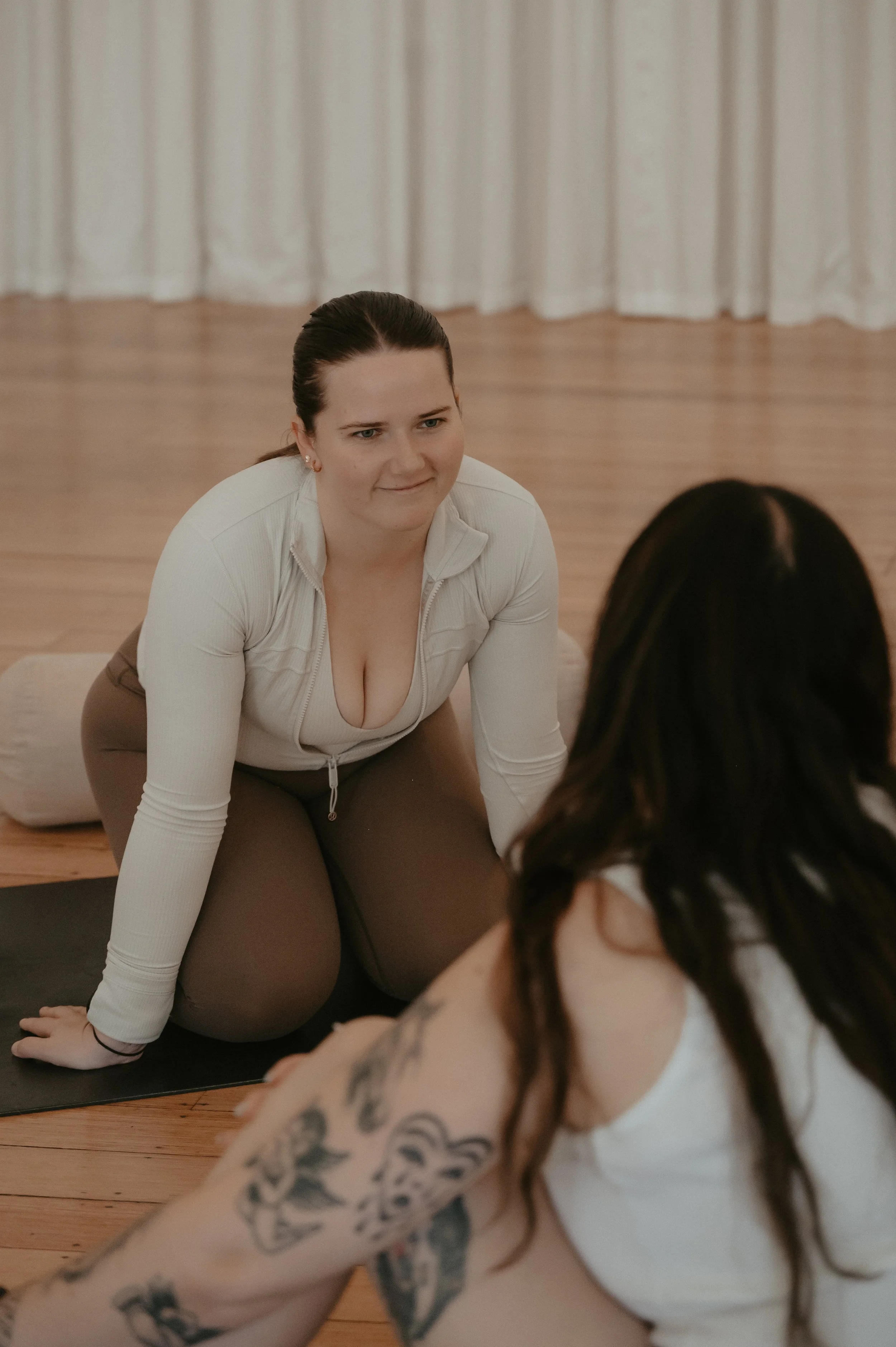 Two women practicing pilates indoors, one with dark hair and tattoos, and the other with dark brown hair in a ponytail, wearing white and brown clothes.