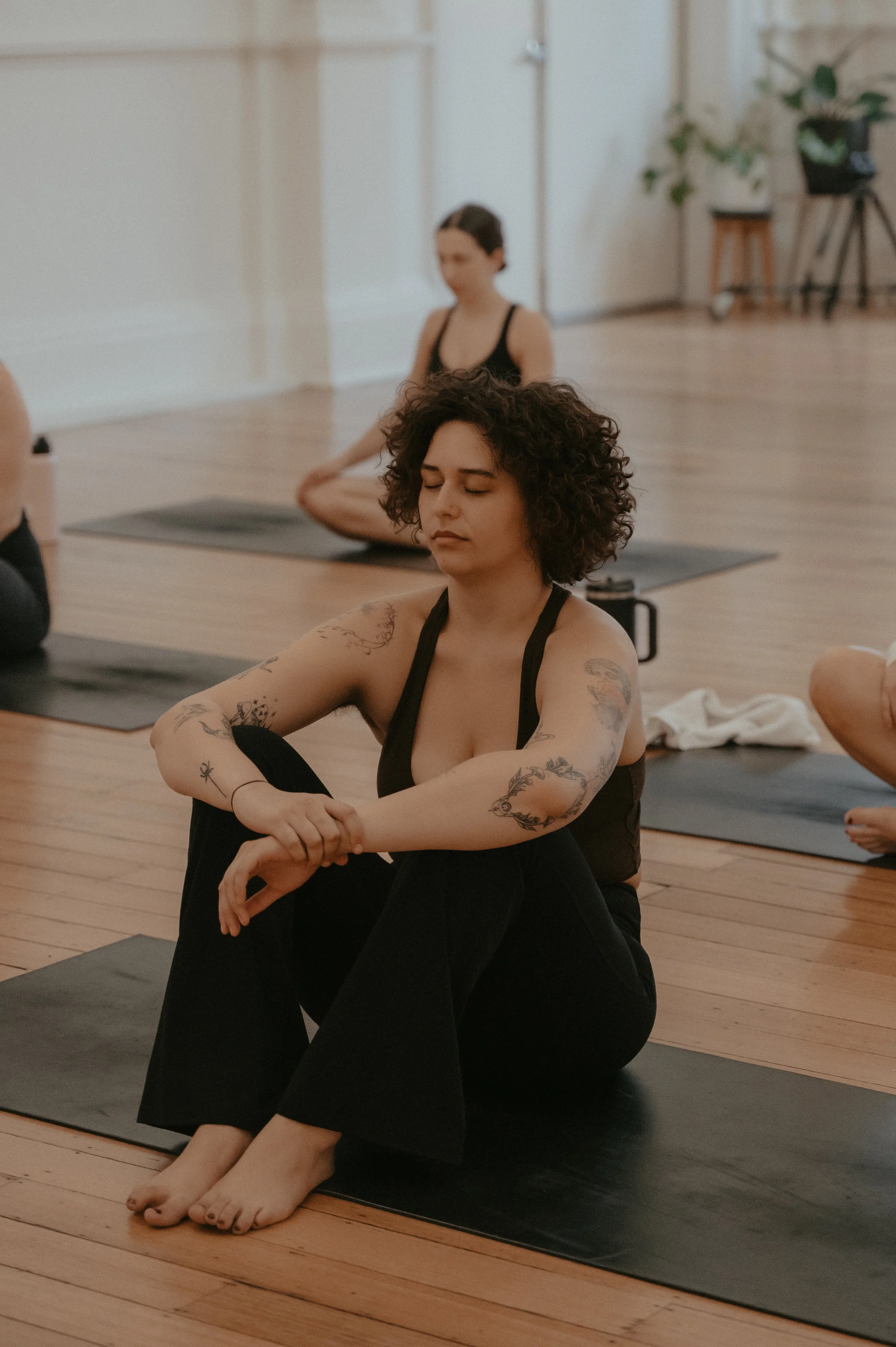 A woman sitting on a pilates mat in a meditation pose with her eyes closed, in a pilates class. Others are practicing pilates in the background.