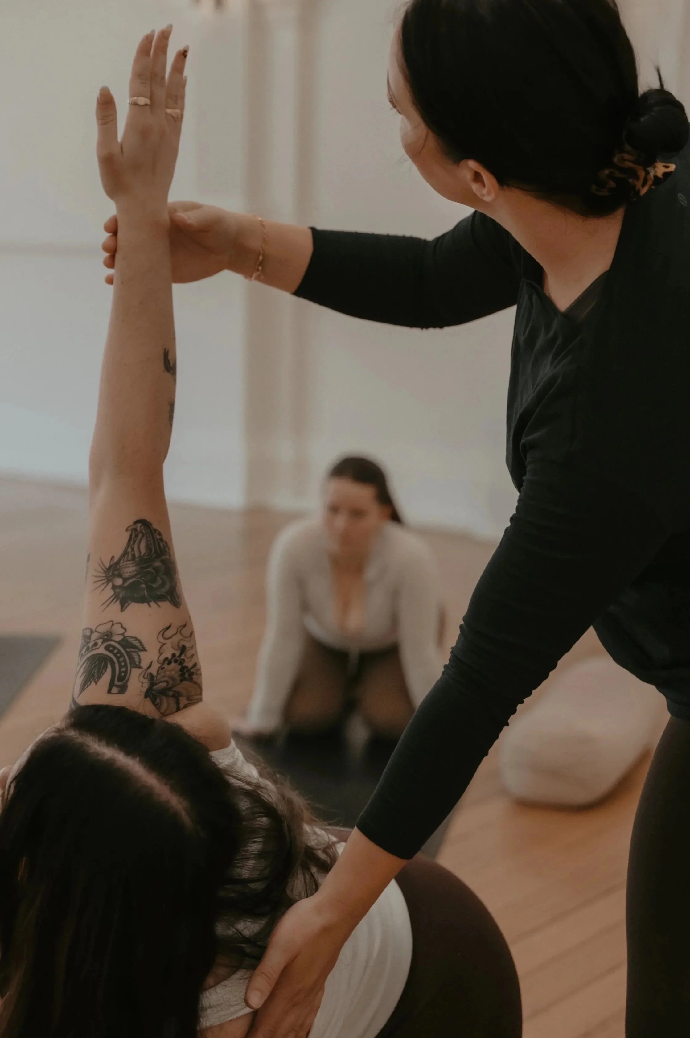 Instructor guiding a woman in a pilates pose during a class, with another woman sitting on the floor observing in the background.