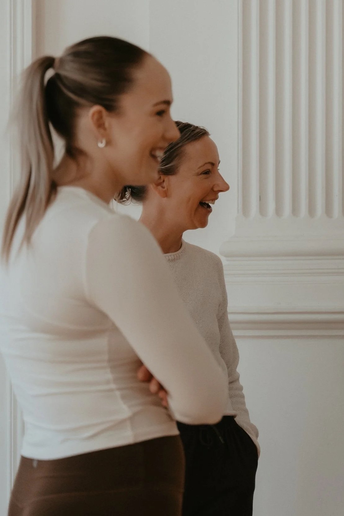 Two women standing indoors, smiling and laughing, with white wall and decorative molding in the background.