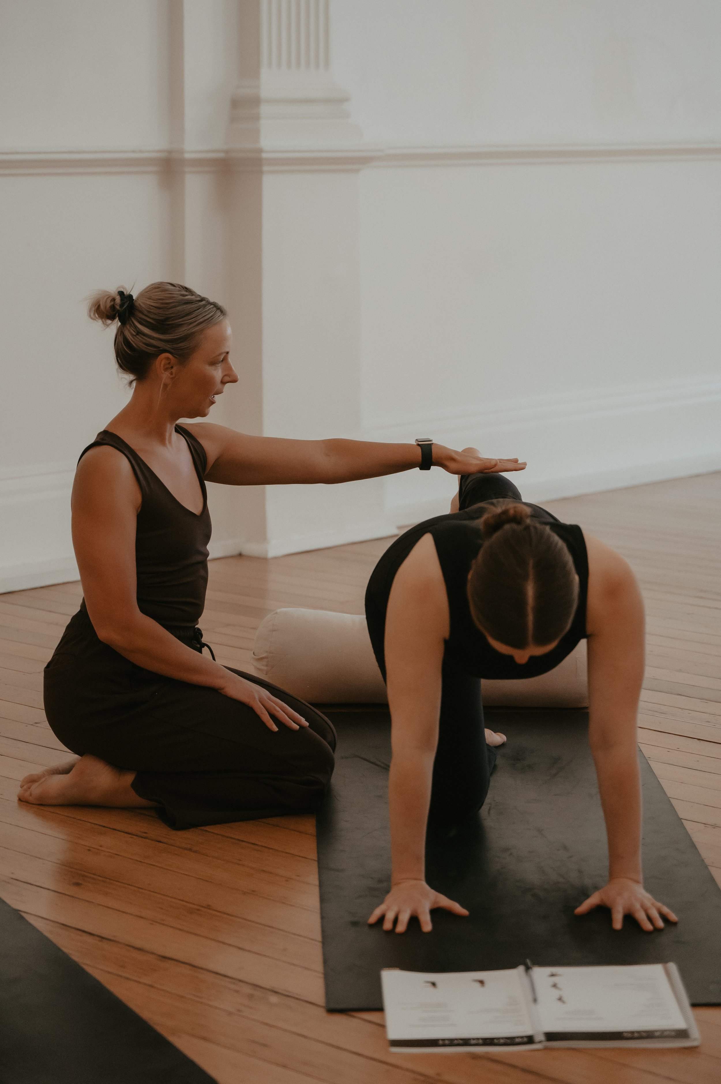 A woman in black is practicing yoga on a black mat, in a plank position with her hands and feet on the ground. A yoga instructor, also in black, kneels beside her, providing guidance with her arm extended and hand on the woman's back. A yoga instruction book or magazine is open on the floor in front of the mat.
