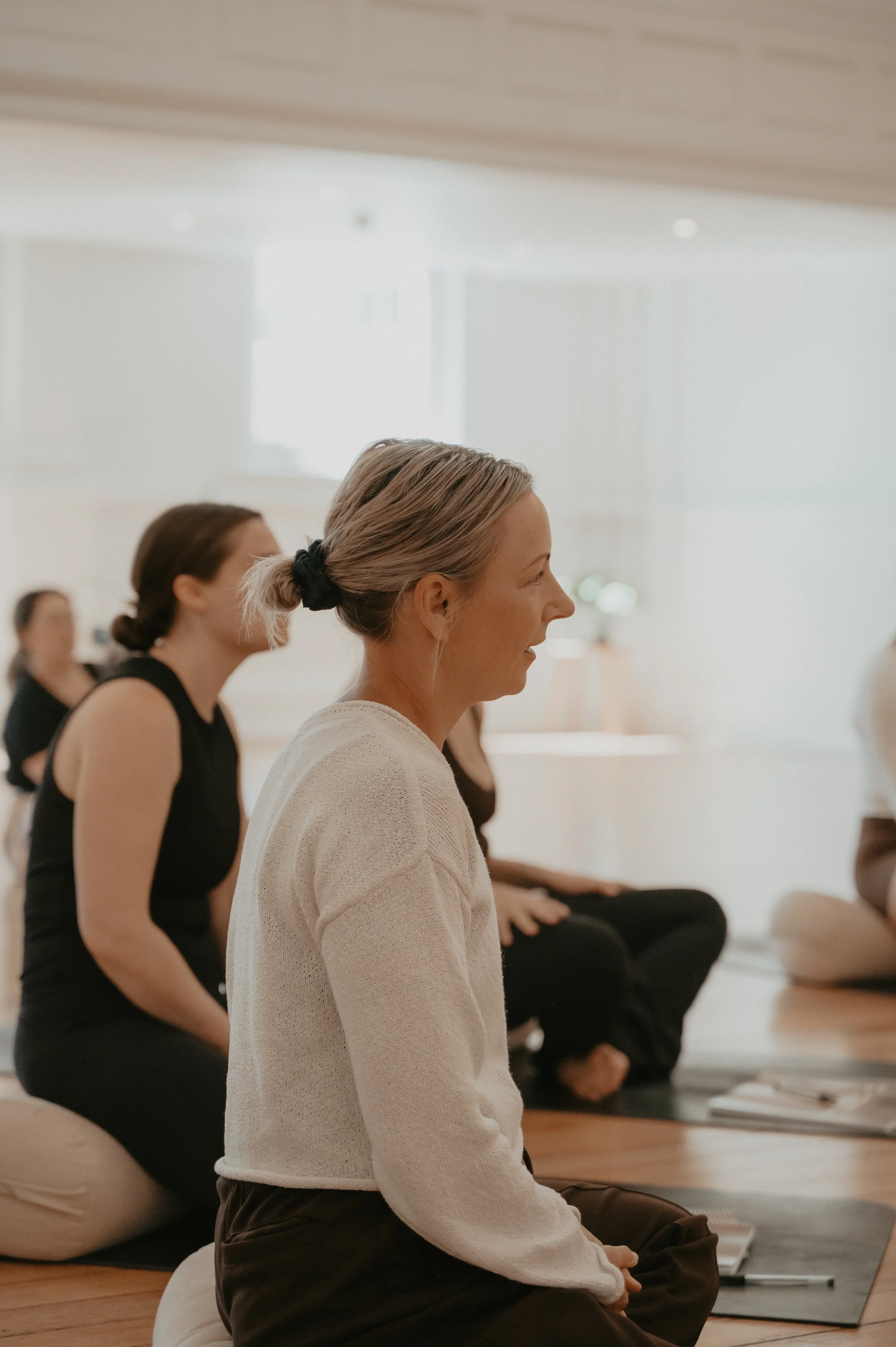 Women sitting in a pilates class learning how to teach pilates, sitting cross-legged on cushions with their eyes closed, in a brightly lit room.