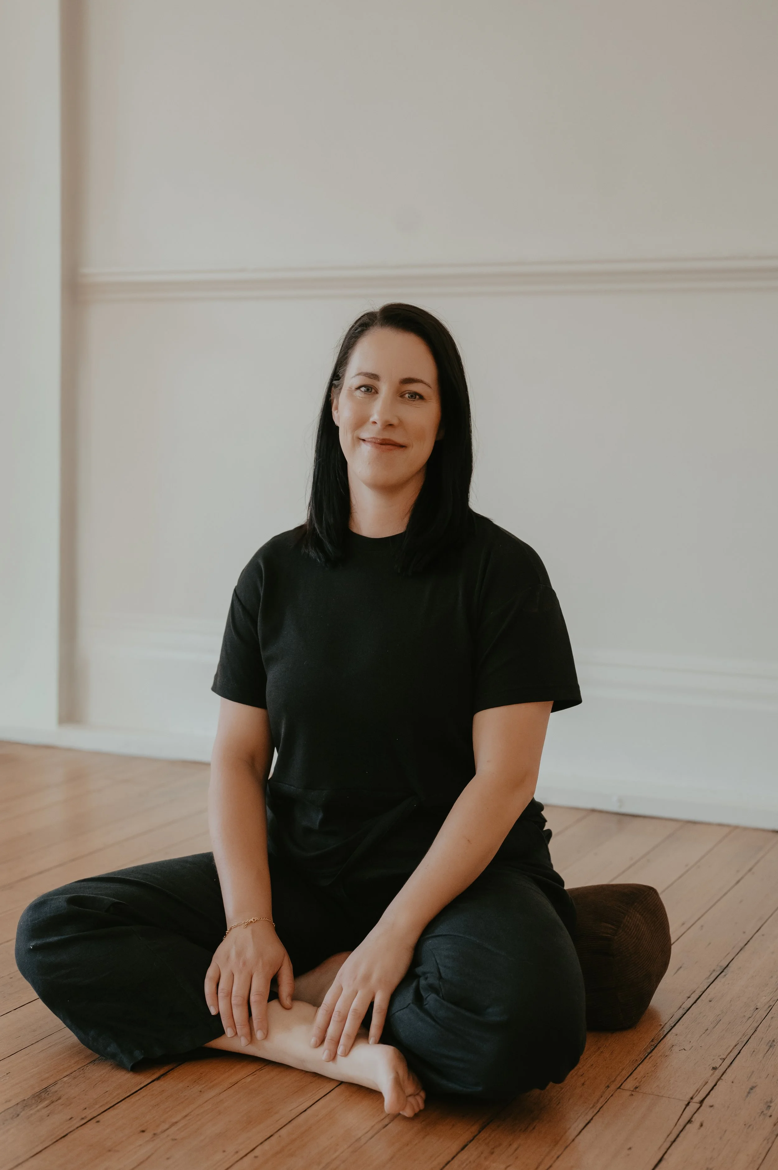 A woman with black hair, wearing a black t-shirt and black pants, sitting on the wooden floor with her legs crossed and smiling softly at the camera in a minimalist room with white walls.