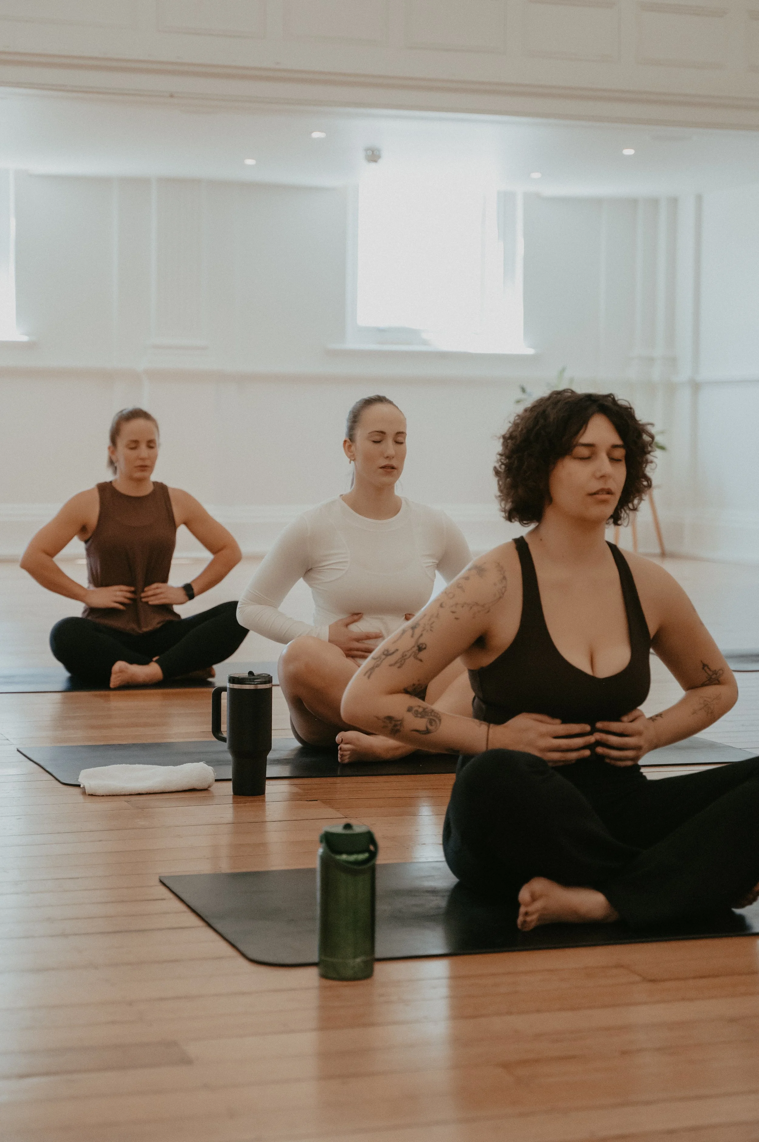 Three women participating in a pilates class, sitting cross-legged on mats with eyes closed, hands resting on their stomachs, in a bright room with wooden floors and large windows.