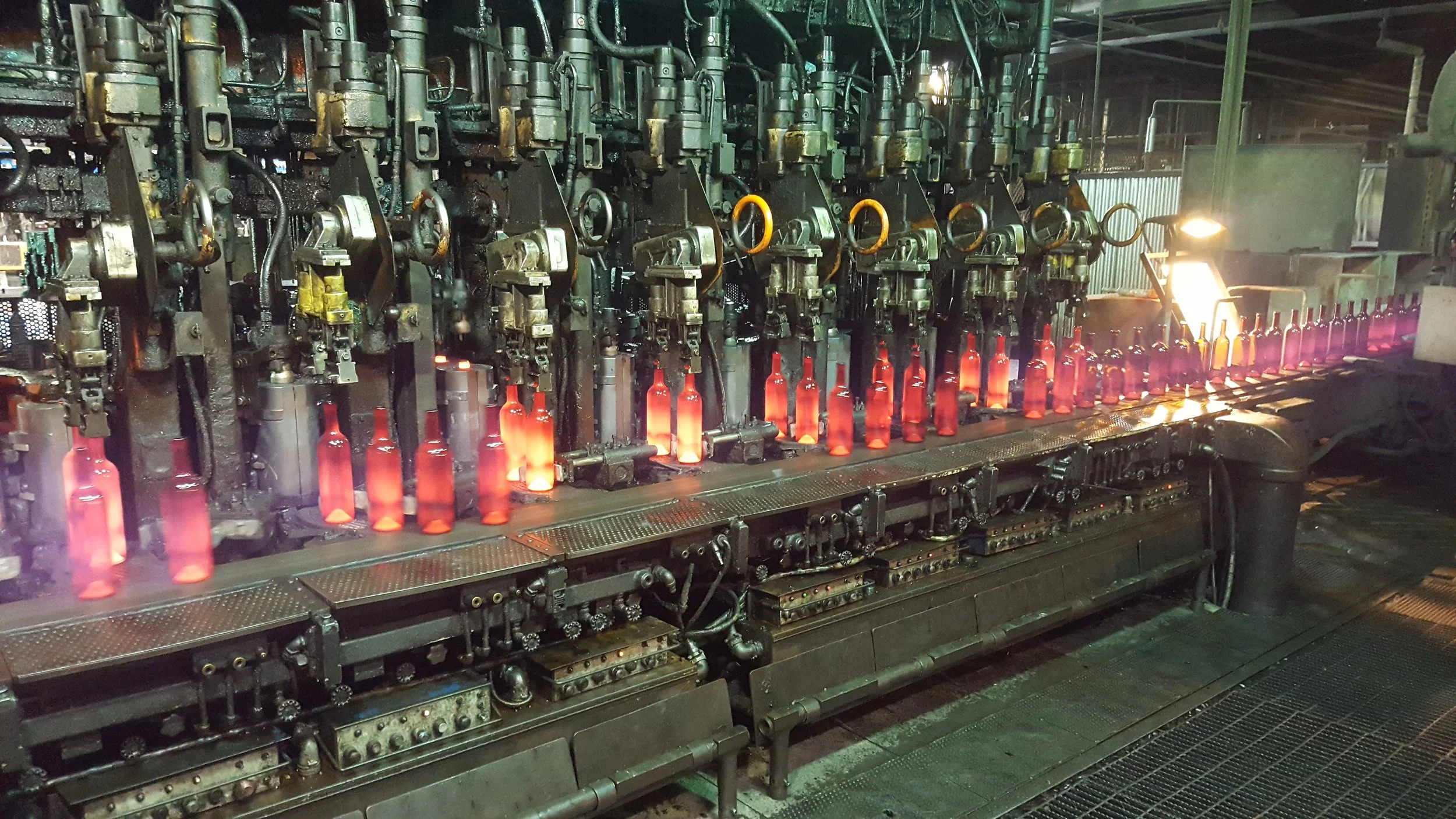 Glass bottles being shaped in a manufacturing factory, with hot, glowing glass and machinery for glass molding. IS style machine producing either beer bottles or wine bottles.