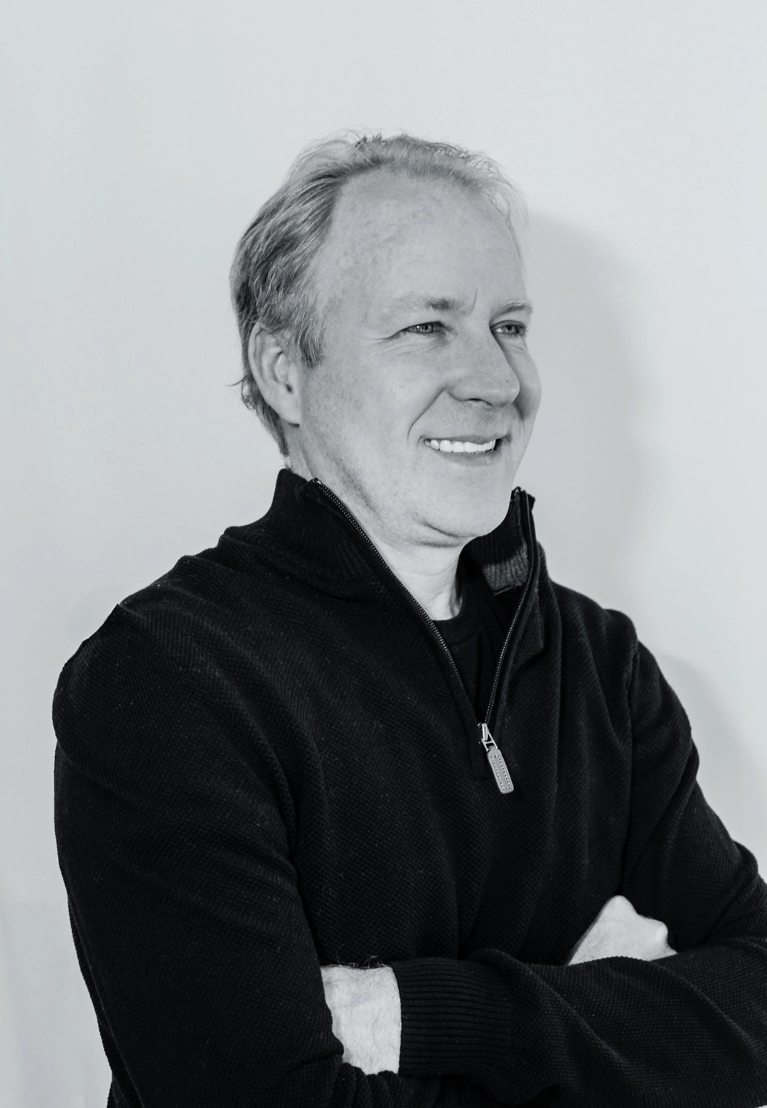 Black-and-white portrait of a smiling man with light hair, wearing a black zip-up sweater, arms crossed, standing against a plain background.