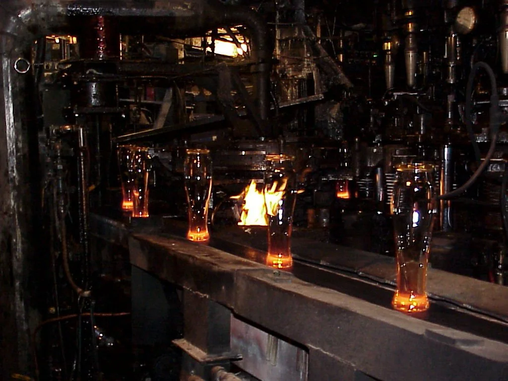 A glass bottle manufacturing process on an assembly line with glowing molten glass bottles being shaped and cooled. H-28  or Harford style machine used to make Tableware either Tumblers, stretch stemware or 2-piece stemware.