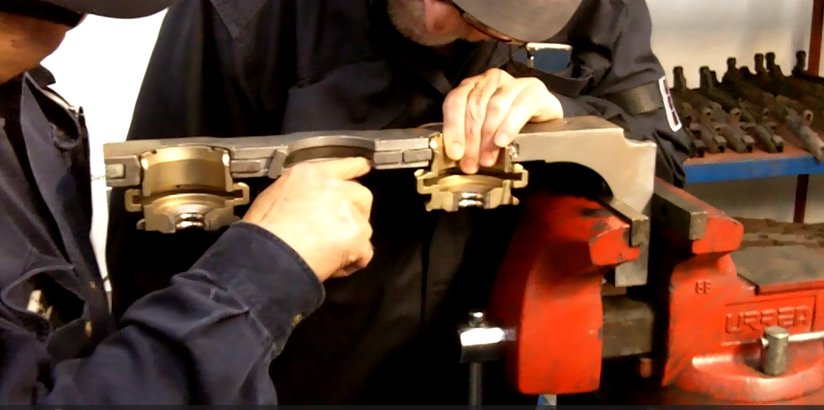 Two workers examining a metal rail mounted on a red vise in a workshop, with tools and equipment in the background.